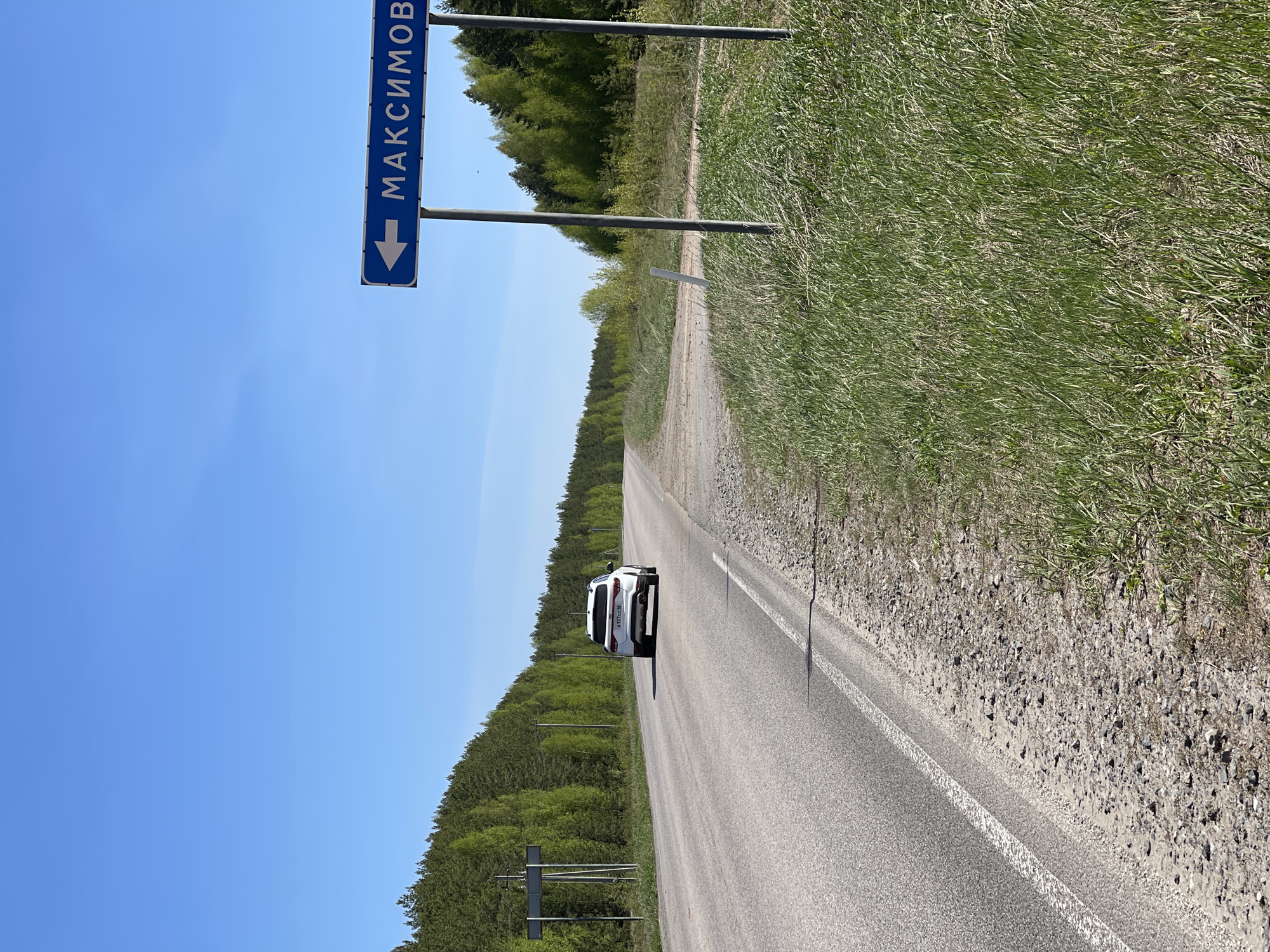 The image depicts a road in Перевозное, Russia. A white car is driving on the asphalt road, which has a white line marking. To the left of the road is a sign pointing to "МАКСИМОВО," and further left is a line of trees. To the right of the road is tall grass. The sky is blue and clear.