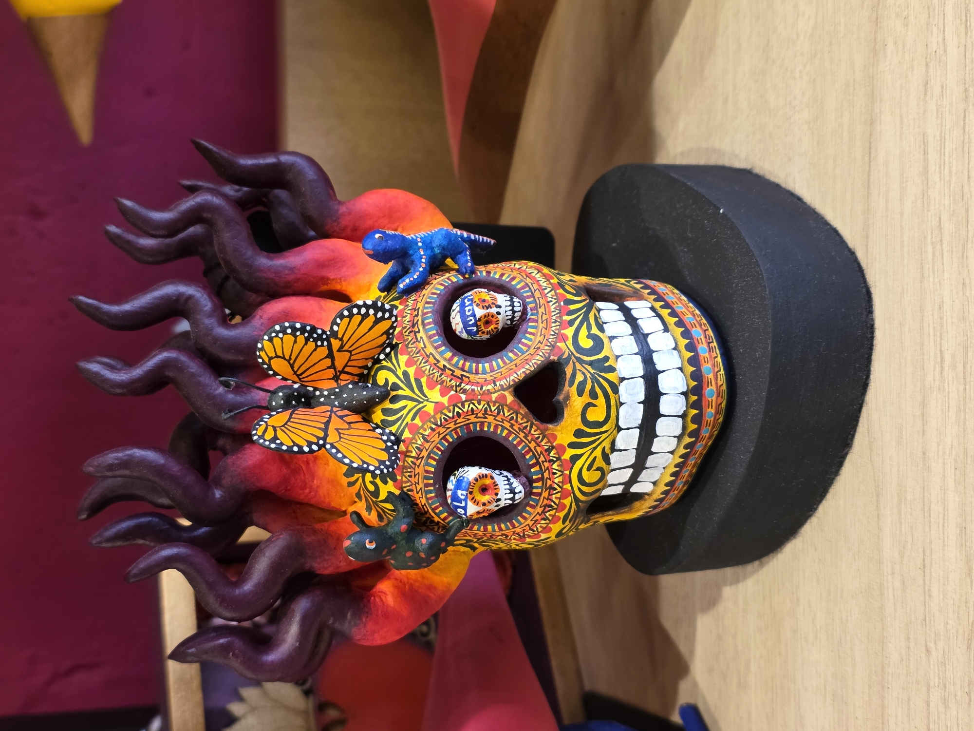 A colorful, hand-painted sugar skull, a traditional Mexican folk art, rests on a dark gray cylindrical stand against a light wood surface. The skull is adorned with intricate designs in vibrant yellows, oranges, and reds. Monarch butterflies and a blue lizard figure are attached to the skull. Within the eye sockets are smaller, decorated skull figurines. The skull also features dark purple, flame-like appendages extending from the back. The scene suggests that the object is located in Puebla, Mexico, based on the distinctive artistic style and symbolism associated with the Day of the Dead celebrations. The precise location within Puebla cannot be determined from the image alone.