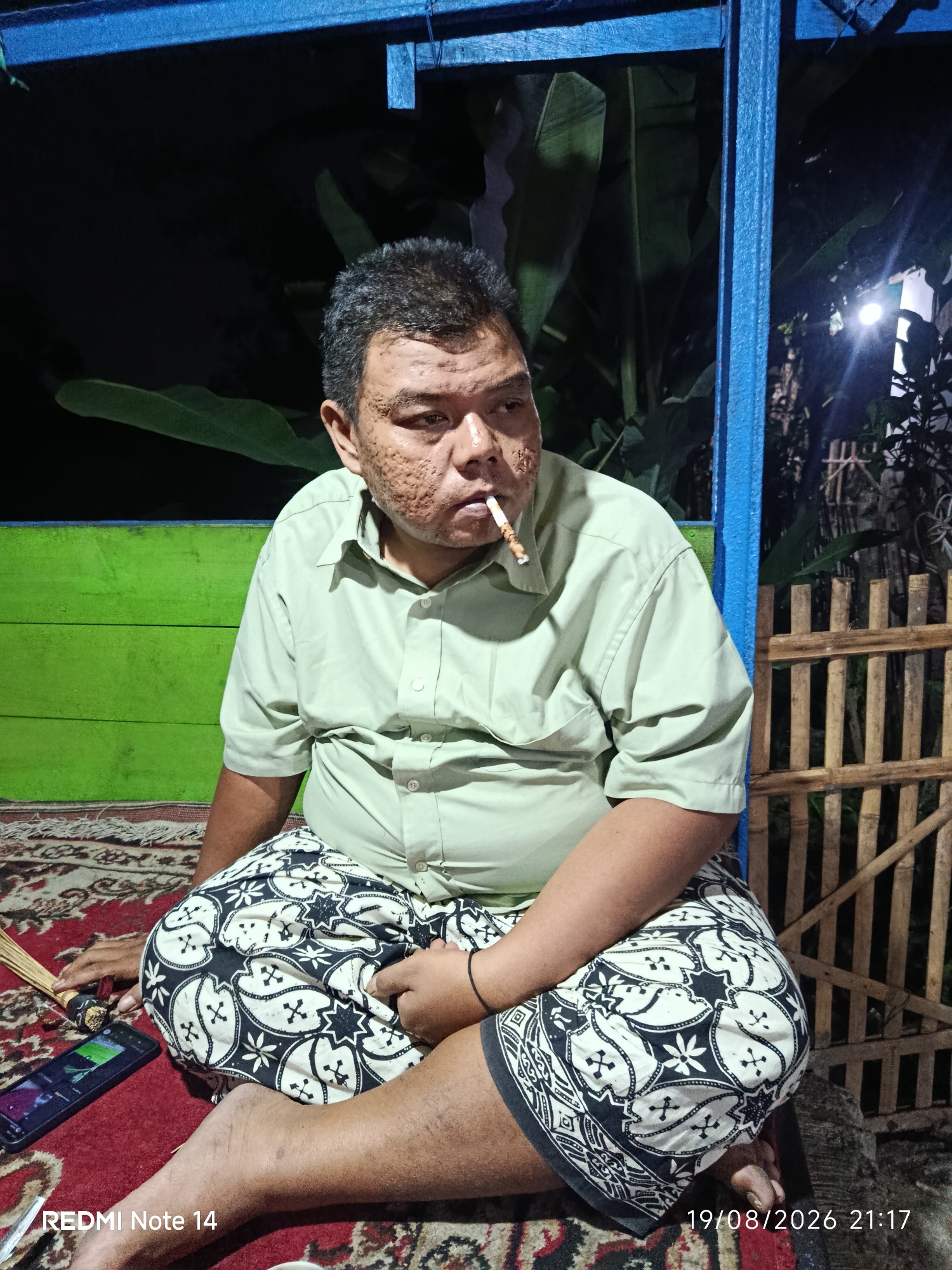 A medium shot captures a man with visible skin texture irregularities sitting cross-legged on a patterned rug. He wears a light green collared shirt and patterned pants. A lit cigarette is held in his mouth. To his left, a cell phone is visible on the rug, and a small broom-like object rests next to it. The background features a green wooden wall and a blue structure overhead. Beyond that, a wooden fence and foliage are dimly lit, suggesting an outdoor evening setting. The timestamp "19/08/2026 21:17" and "REDMI Note 14" are present in the lower right and left corners, respectively, indicating the image was taken on August 19, 2026, at 9:17 PM, using a Redmi Note 14 device. The location is identified as Panggungrejo, Indonesia.
