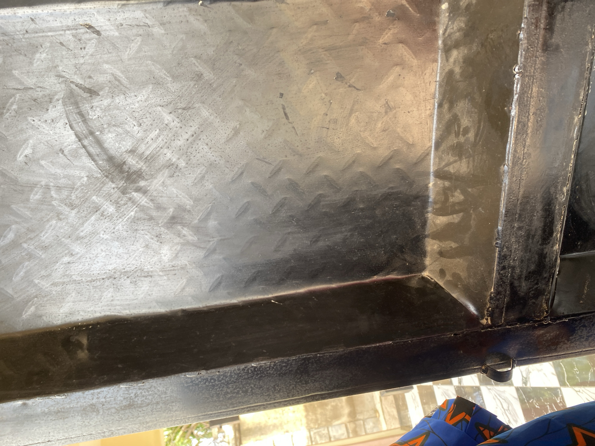 The image shows a close-up of a metal surface, likely part of a gate or structure, with a diamond plate pattern. The metal appears weathered and shows signs of use, with scratches and smudges. A portion of a dark, vertical metal beam is visible on the left side, and a curved handle is attached to it. In the lower left corner, a person's leg is visible, wearing a blue fabric with an orange and black pattern, suggesting it is clothing. The background, visible through a gap on the left, shows a paved area and some greenery, indicating an outdoor setting. The overall scene is a forensic description of an object and its immediate surroundings. There is no visible indication that this scene is located in Jauro Yinu, Nigeria.