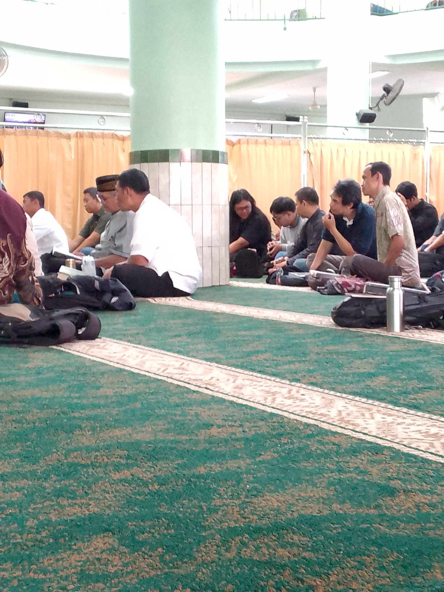 In Cimahi, Indonesia, a group of men are seated on a green carpeted floor, likely inside a mosque or prayer hall, as indicated by the architectural elements like the column and beige curtains that section off areas. They are dressed in casual attire, with some wearing white shirts and others in muted colors. Several individuals have bags and a water bottle placed on the carpet near them. The men appear to be engaged in a period of rest or contemplation. The lighting suggests it is daytime, possibly after a prayer service. The overall scene depicts a communal gathering, emphasizing a sense of quietude and shared space.