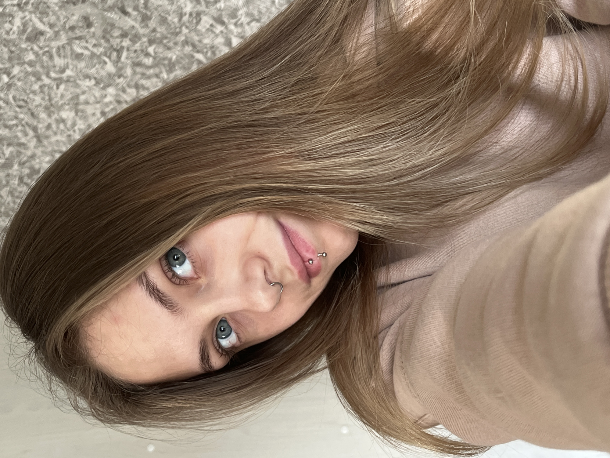 A young woman with light brown hair and blue eyes is taking a selfie. She has facial piercings on her nose and lip. She is wearing a light pink sweatshirt. The background is out of focus but appears to be a carpeted floor. The image is rotated slightly to the left. There is no indication of the location being Фоки, Russia.
