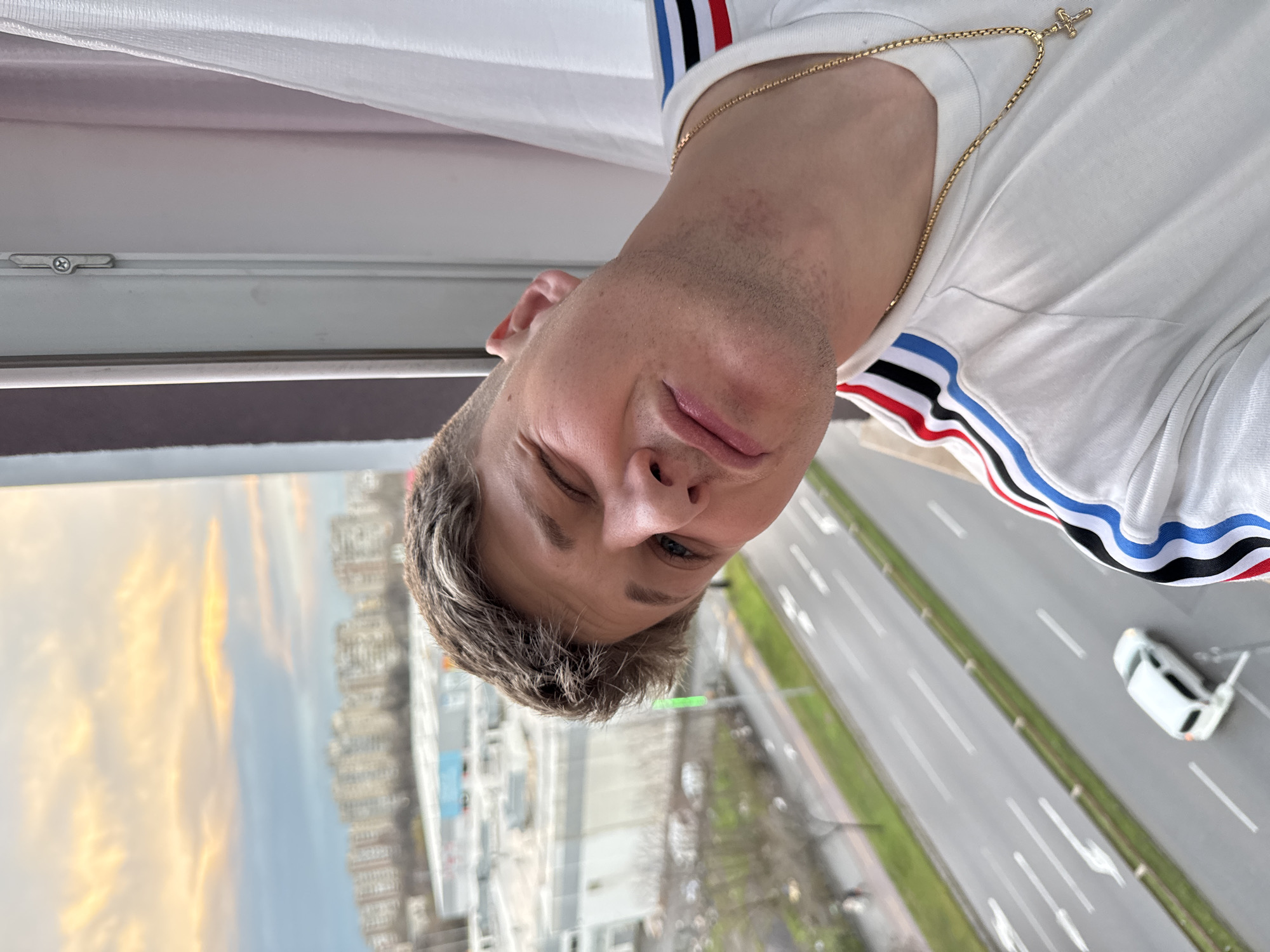 A close-up, portrait-style shot, is oriented sideways, depicting a fair-skinned man wearing a white t-shirt with red, black, and blue stripes along the shoulder. He has short, light-colored hair with visible stubble on his face, and is wearing a gold necklace with a cross pendant.
Outside the window, a multi-lane highway with traffic including a white car is visible, along with city buildings and a sky tinted by sunset colors. The scene suggests an elevated view, potentially from a high-rise building, in Plovdiv, Bulgaria.