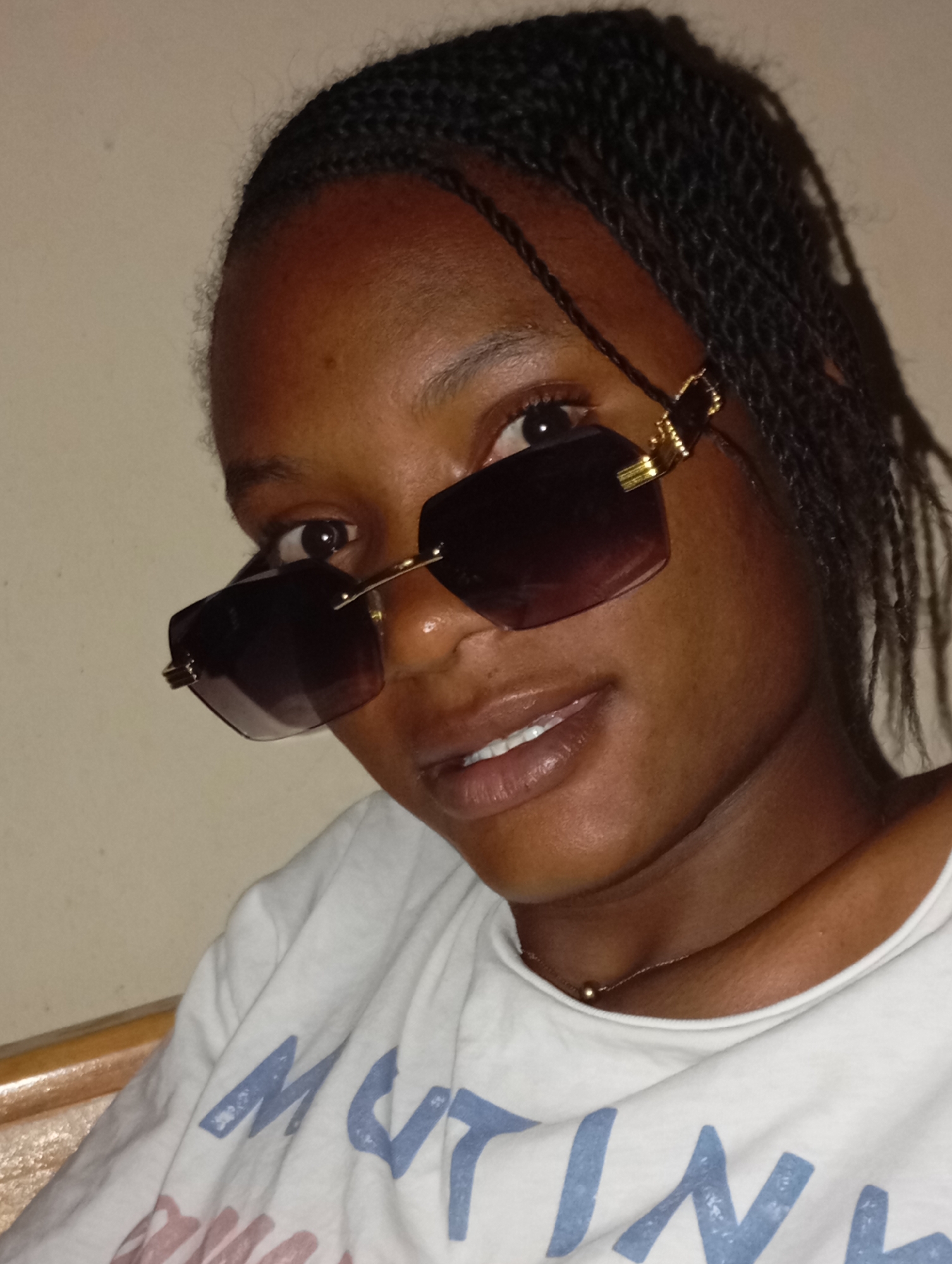 A close-up shot shows a young, dark-skinned woman wearing braided hair and sunglasses. She is looking towards the camera with a slight smile. She is wearing a white t-shirt and a delicate gold necklace. The background appears to be a plain, light-colored wall. The location is Unknown, Unknown.