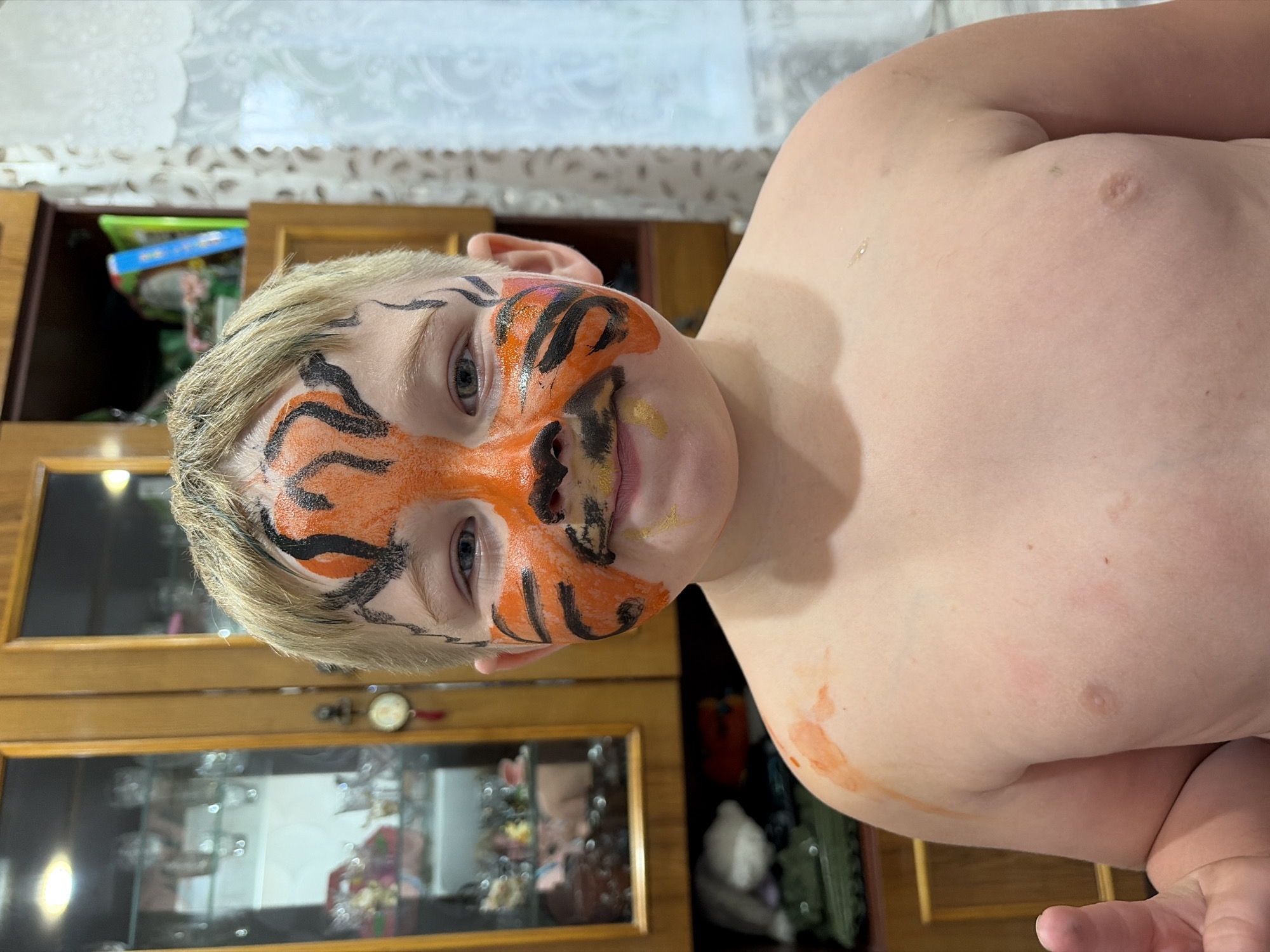 The image shows a young child with tiger face paint on. The child is looking towards the camera, with their head tilted to the side. The face paint is orange and black, with black stripes and markings around the eyes and nose. The child has blonde hair.
The child is in an indoor setting, with a wooden cabinet visible behind them. The cabinet has glass doors, and some items are visible inside. The lighting suggests it is daytime.
The child appears to be relaxed and comfortable. The overall scene is casual and domestic. There is no visible text in the image.