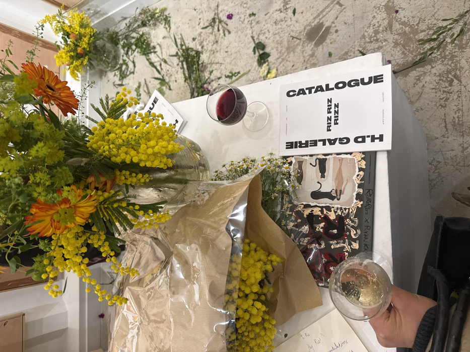 A brightly lit, high-angle shot shows a table laden with flowers, catalogues, and wine glasses. A large bouquet of yellow mimosa and orange gerbera daisies dominates the left. A brown paper-wrapped bunch of mimosa sits center. A glass of red wine and a white "Catalogue" with "H.D. Galerie" text are visible. A book featuring cat illustrations rests near a partially visible card. A hand holding a glass of white wine appears in the lower right. Sprinkled flower petals and greenery scatter on the table and floor.