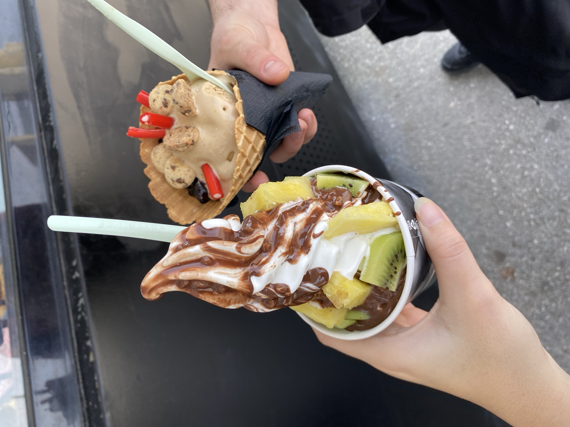 A close-up, high-angle shot captures two people holding different desserts outdoors. On the left, a hand, likely masculine, holds a waffle cone filled with a light brown, creamy dessert, adorned with cookie pieces and several red stick-like candies. A black napkin is wrapped around the cone, and a light green plastic spoon is inserted into the treat.
On the right, another hand, likely feminine, holds a white paper cup containing a generous swirl of white soft-serve dessert, heavily drizzled with chocolate sauce. This cup is topped with vibrant chunks of yellow pineapple and green kiwi. A matching light green plastic spoon, faintly inscribed with "Frozen Yogurt," is also present in this dessert.
The background on the left features a dark, reflective surface, possibly a car hood or a dark table. To the right, gray concrete or pavement is visible, along with parts of a person's black clothing and shoe. The natural lighting indicates it is daytime. The overall scene suggests a casual and enjoyable moment of sharing sweet treats in an outdoor setting. No specific location cues are visible within the image itself.