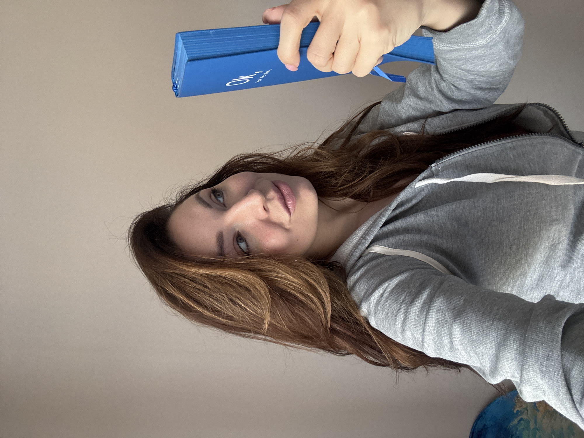 A young woman holds a blue, rectangular object in her left hand. She is wearing a gray hooded sweatshirt with white drawstrings. She is looking directly at the camera. Her hair is light brown. The background is a solid, pale wall. A blue and green object is visible in the lower right corner of the frame. The image is slightly rotated counter-clockwise. The location is Unknown, Unknown.