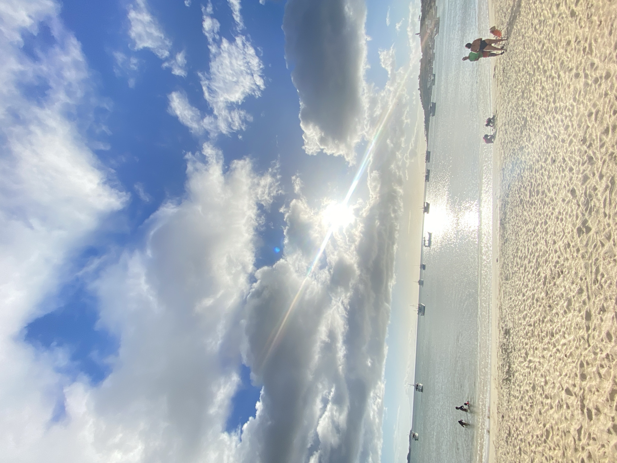 The image shows a beach scene in Juan Griego, Venezuela. The sky is blue with white and gray clouds, with the sun shining brightly behind some of them. Sunlight reflects off the water, creating a glare. Several boats are anchored in the bay. On the sandy beach, several people are visible. Some stand near the water's edge, while others appear to be playing in the sand or shallow water. The sand is light-colored, and the patterns of footprints and ripples are visible.