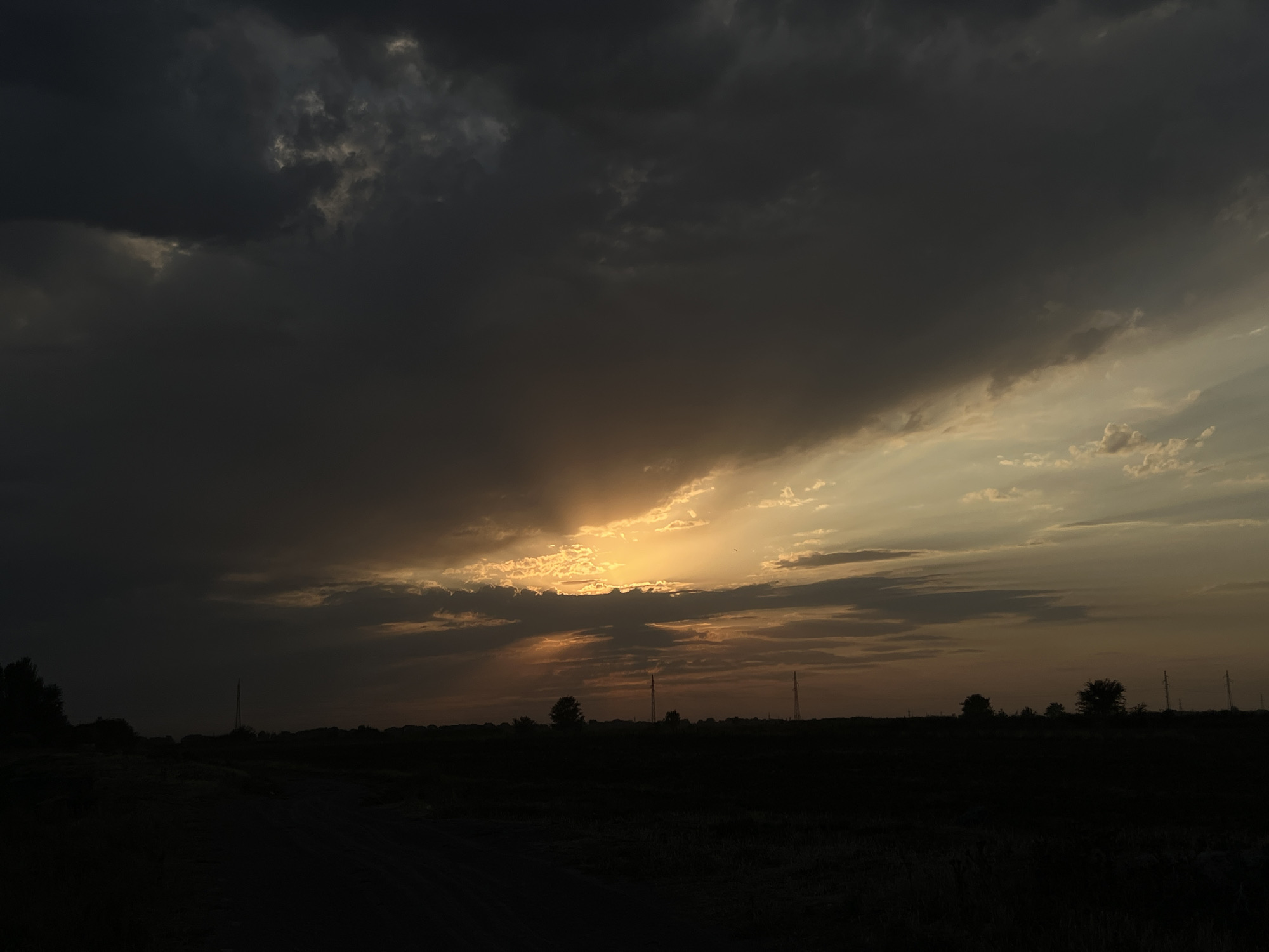 This is a photograph of a dark, cloudy sunset over a rural landscape. The sky is dominated by heavy, dark clouds, with a sliver of bright orange and yellow light breaking through near the horizon, suggesting the sun is setting. Below the sky, a silhouetted landscape of dark fields and vegetation is visible, with a dirt road or path leading into the foreground. Several tall, thin electrical towers are scattered across the horizon, their dark forms contrasting with the sky. There are also silhouettes of trees and what appear to be low-lying structures in the distance. The overall impression is one of a dramatic and somewhat somber sky over a quiet, open rural area. The location is identified as Дачное, Kyrgyzstan.