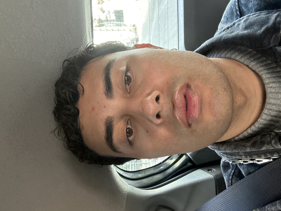 The image presents a close-up portrait of an adult individual, appearing male, seated within the rear passenger compartment of a vehicle in Neuilly-Plaisance, France. The subject has a medium skin tone, dark curly hair, and brown eyes, with a small red blemish on the forehead. The lips are full and slightly parted, exhibiting a glossy appearance. A small hoop earring is visible in the subject's right ear. The individual is dressed in a grey ribbed turtleneck sweater beneath a dark wash denim jacket. A silver-colored chain with multiple links is visible across the left side of the subject's chest, secured to the jacket. A dark blue seatbelt strap is diagonally positioned across the left shoulder and chest. The subject maintains a direct gaze towards the camera with a neutral expression. The vehicle's interior is visible, featuring a grey headliner and grey plastic trim of the rear left door. Through the vehicle's windows, an outdoor daylight scene is discernible, depicting a street with blurred buildings, vehicles, and trees, indicative of an urban or suburban setting.