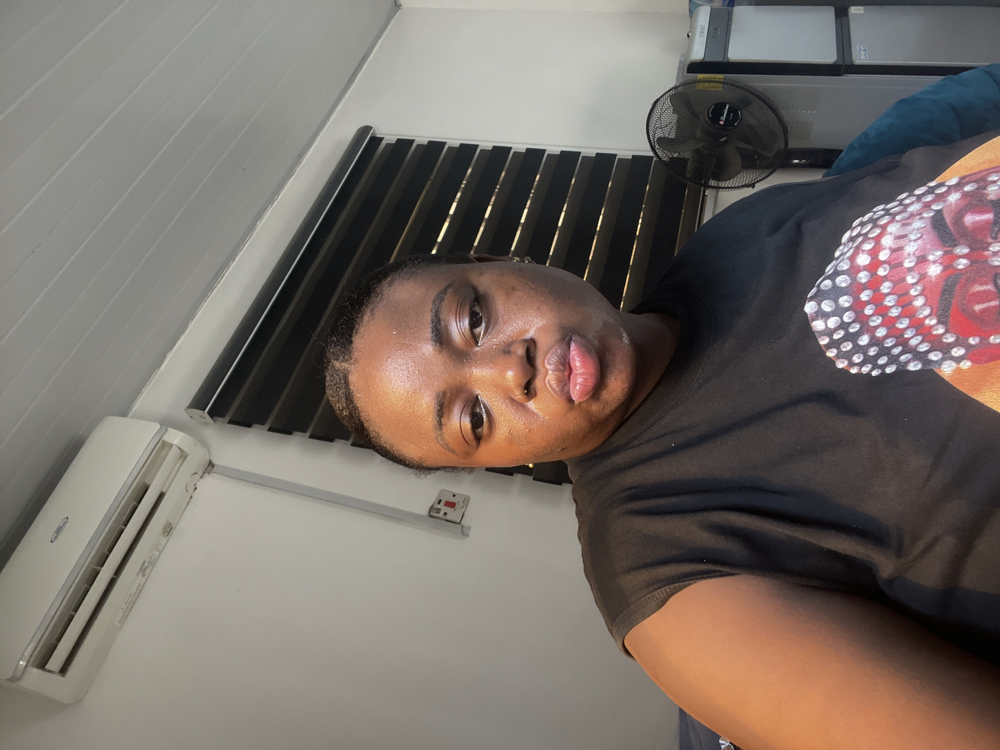 A close-up, angled indoor shot focuses on a woman wearing a black t-shirt with a colorful design on the front. Her face is prominent, and she appears to be looking directly at the camera. Behind her, a black window blind and an air conditioning unit mounted on the wall are visible. The room appears to be in a residential setting. Given the request, the location is in the city of Abuja, Nigeria. A standing black fan is also in the background.