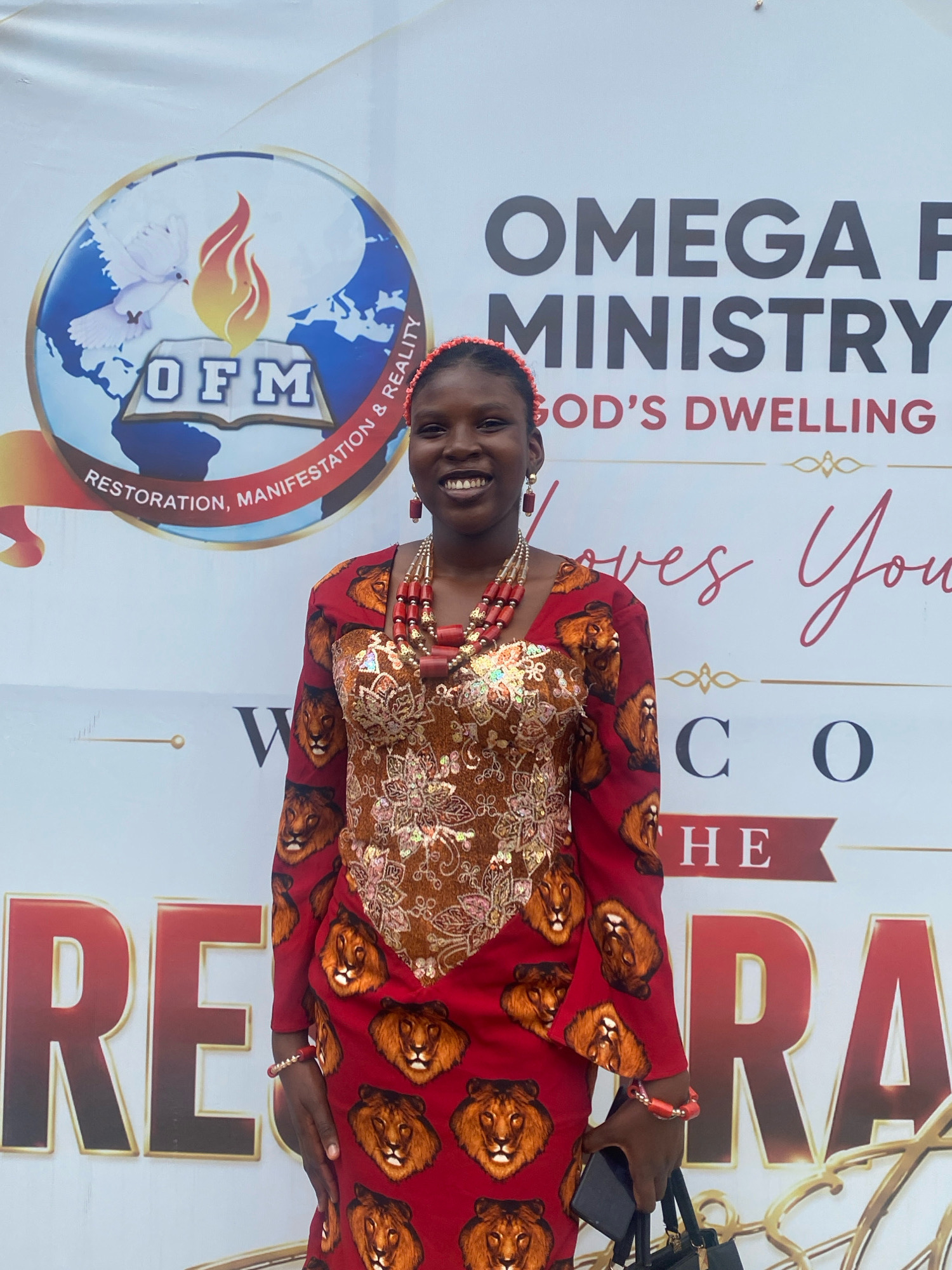 A young Black woman stands in front of a banner that reads "OMEGA F MINISTRY GOD'S DWELLING LOVES YOU THE RE RA". The woman wears a red dress with a lion print and a beaded necklace. She smiles at the camera. The banner appears to be in Ado, Nigeria.