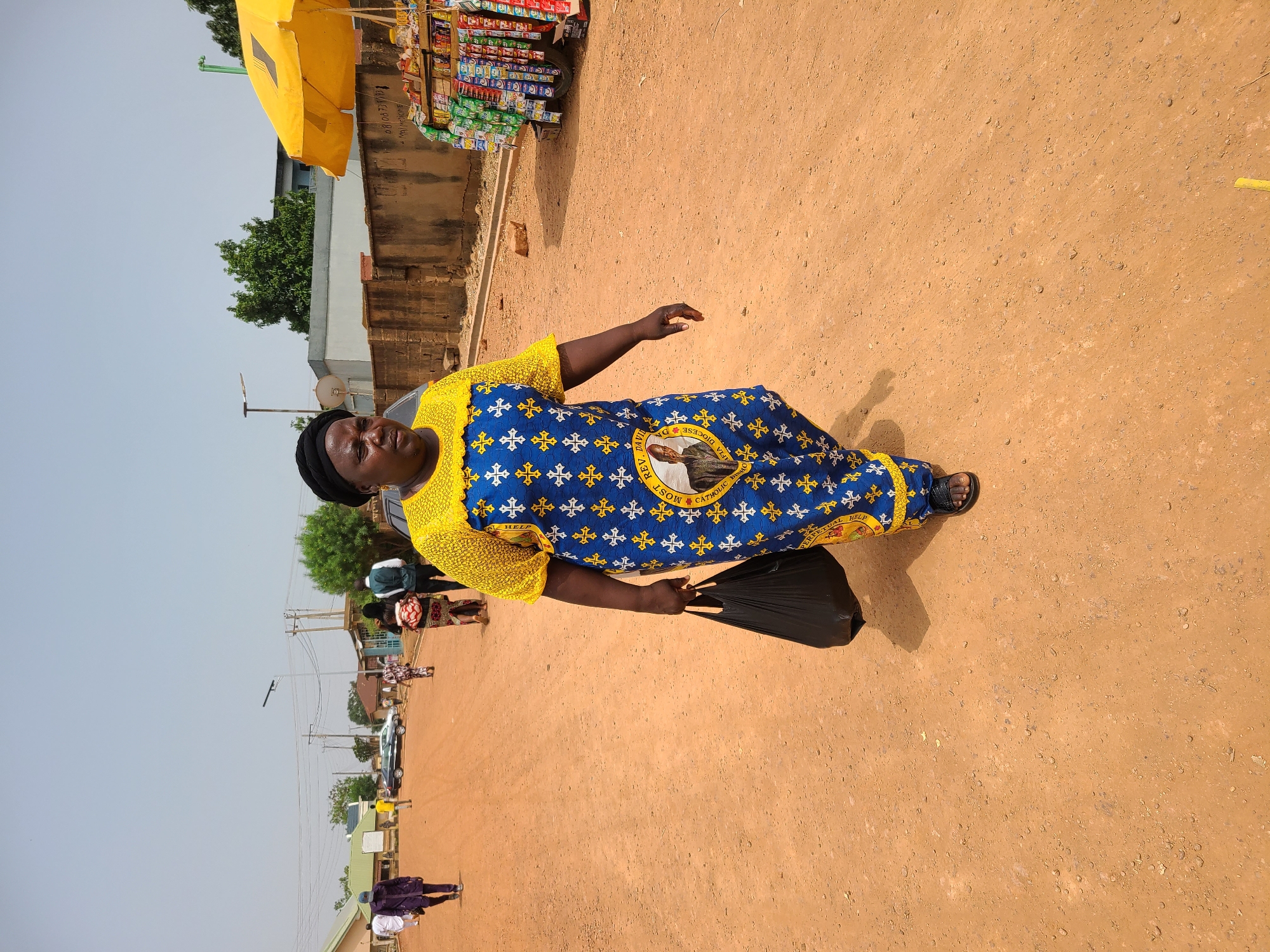 An adult woman stands on a reddish-brown, dirt-covered street. She wears a patterned blue and yellow dress and black head covering, and carries a black plastic bag. Behind her is a street scene with buildings, trees, utility poles, and other pedestrians. A stall with a yellow umbrella and various snacks is to her right. The location is Jos, Nigeria.