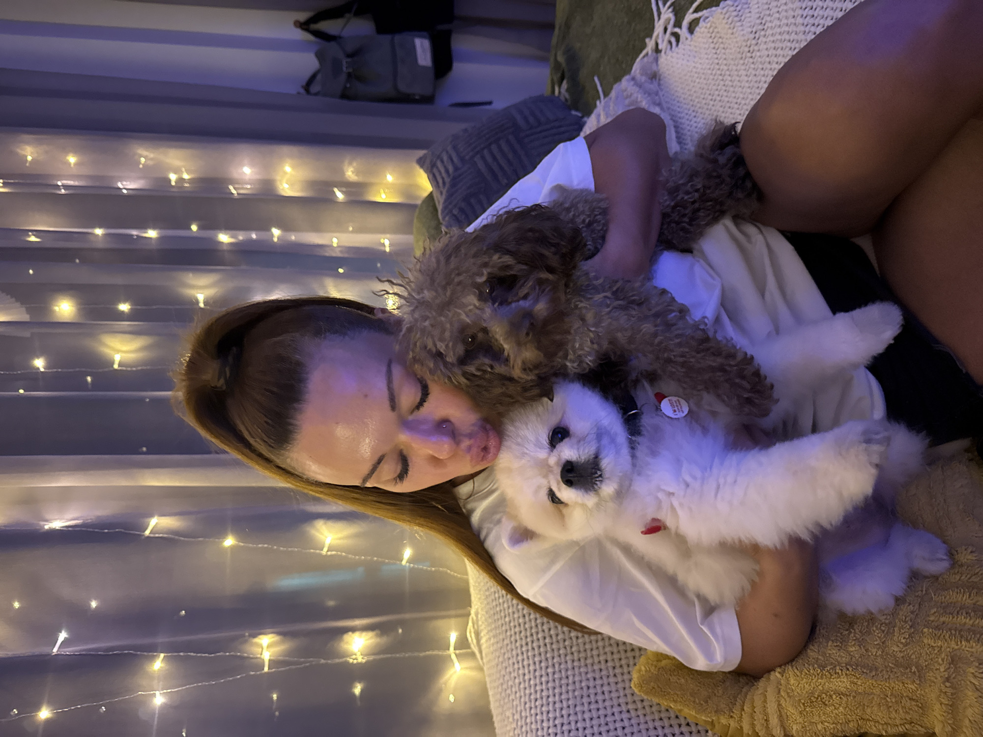 In Thep Krasatti, Thailand, a woman with her hair pulled back in a high ponytail is seated on a couch, holding two small dogs. One dog, a white Pomeranian, is nestled in her arms, while a brown poodle-like dog is held close on her right. The woman is leaning in, appearing to kiss the Pomeranian. She is wearing a white t-shirt. Behind her, sheer white curtains are adorned with a string of warm-toned fairy lights, creating a cozy atmosphere. A backpack hangs on a hook on the wall in the background. The scene is illuminated by the soft glow of the lights and ambient room lighting.