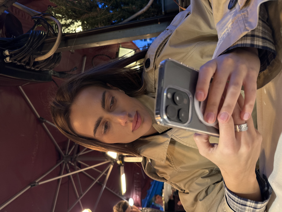 A young adult female is depicted from the chest up, seated at a table in an outdoor or semi-outdoor restaurant setting in Venice, Italy. She has long, dark brown hair and is wearing a light tan trench coat over a plaid shirt, the cuff of which is visible on her left arm. A silver-toned ring with circular motifs is present on her left ring finger. She is looking downwards, focused on a gray smartphone held in both hands, which is encased in a clear protective case and has a white circular accessory attached to its back. The phone's multi-lens camera array is visible. Overhead, a large, dark red umbrella canopy with visible structural ribs and internal lighting fixtures covers the area. In the blurred background to the left, another individual is partially visible. The upper right background shows bundled electrical cables and green foliage. The table in the foreground is covered with a white tablecloth.