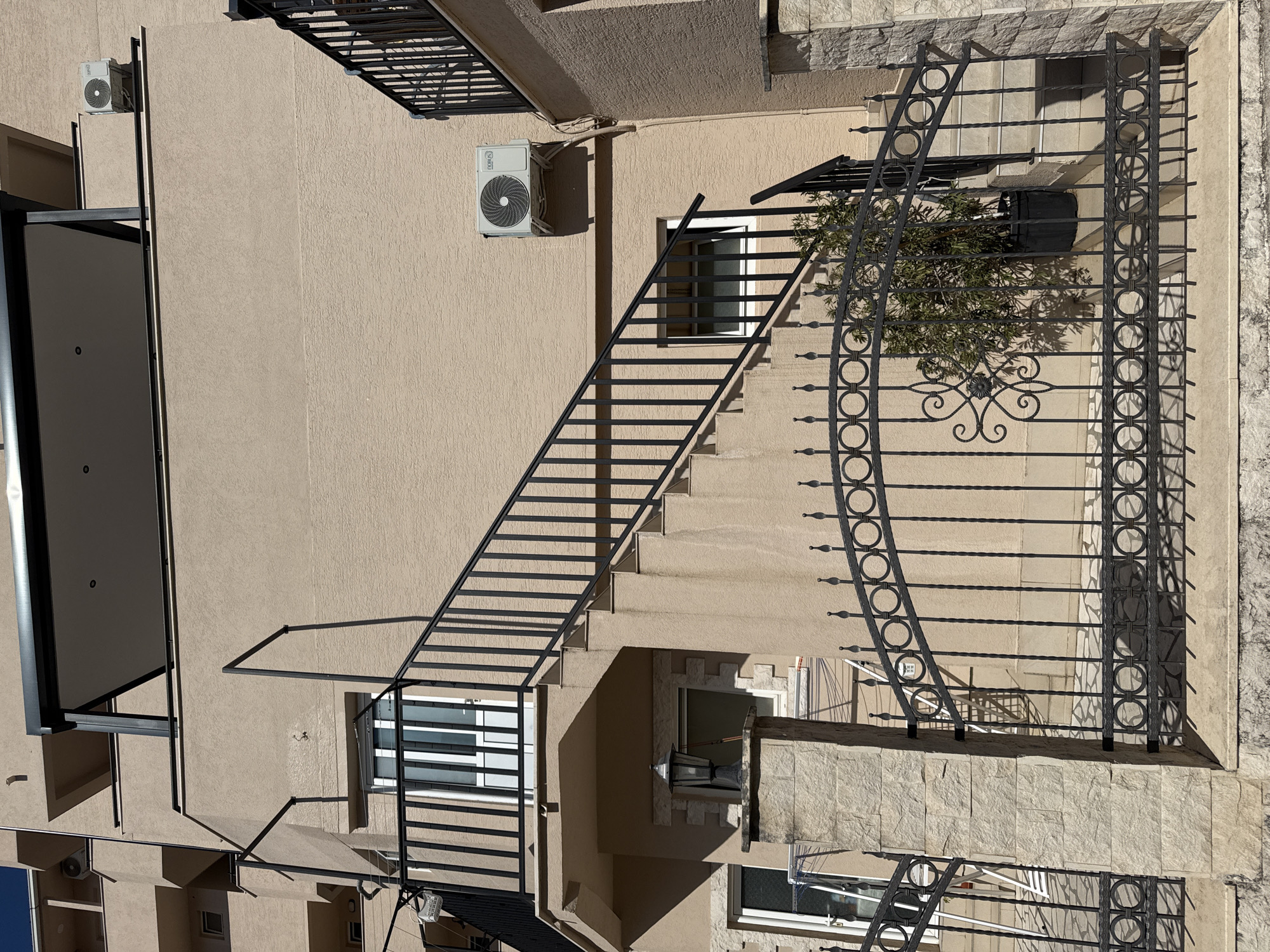 The image displays the exterior facade of a multi-story building, likely an apartment complex or hotel, characterized by a light beige stucco finish and sections of light-colored stone cladding, particularly visible near the base and around windows.
The most prominent feature is a black metal staircase, extending diagonally upwards across the frame. This staircase features numerous horizontal bars and casts strong, well-defined shadows onto the textured wall, indicating bright daylight and clear skies, likely mid-morning or early afternoon. Ornate black metal railings, decorated with circular and scroll patterns, are present on what appear to be balconies or lower-level walkways.
Several windows are visible, some open, and others featuring internal horizontal blinds or external security grates. Two white outdoor air conditioning units are mounted on the wall; the brand "GREE" is clearly visible on one of them. A small green leafy plant or bush grows near one of the ornate railings, also casting a shadow. An exterior wall light fixture is affixed to the building near a window. In the upper left corner, a modern black metal canopy or awning structure extends from the facade.
No people are present, and no specific activity or event is taking place, presenting a static architectural scene. The overall aesthetic and materials suggest a setting consistent with the Mediterranean or Southern European region, aligning with the context of Budva, Montenegro. The perspective appears to be from an elevated position, looking across and slightly down at the building.
