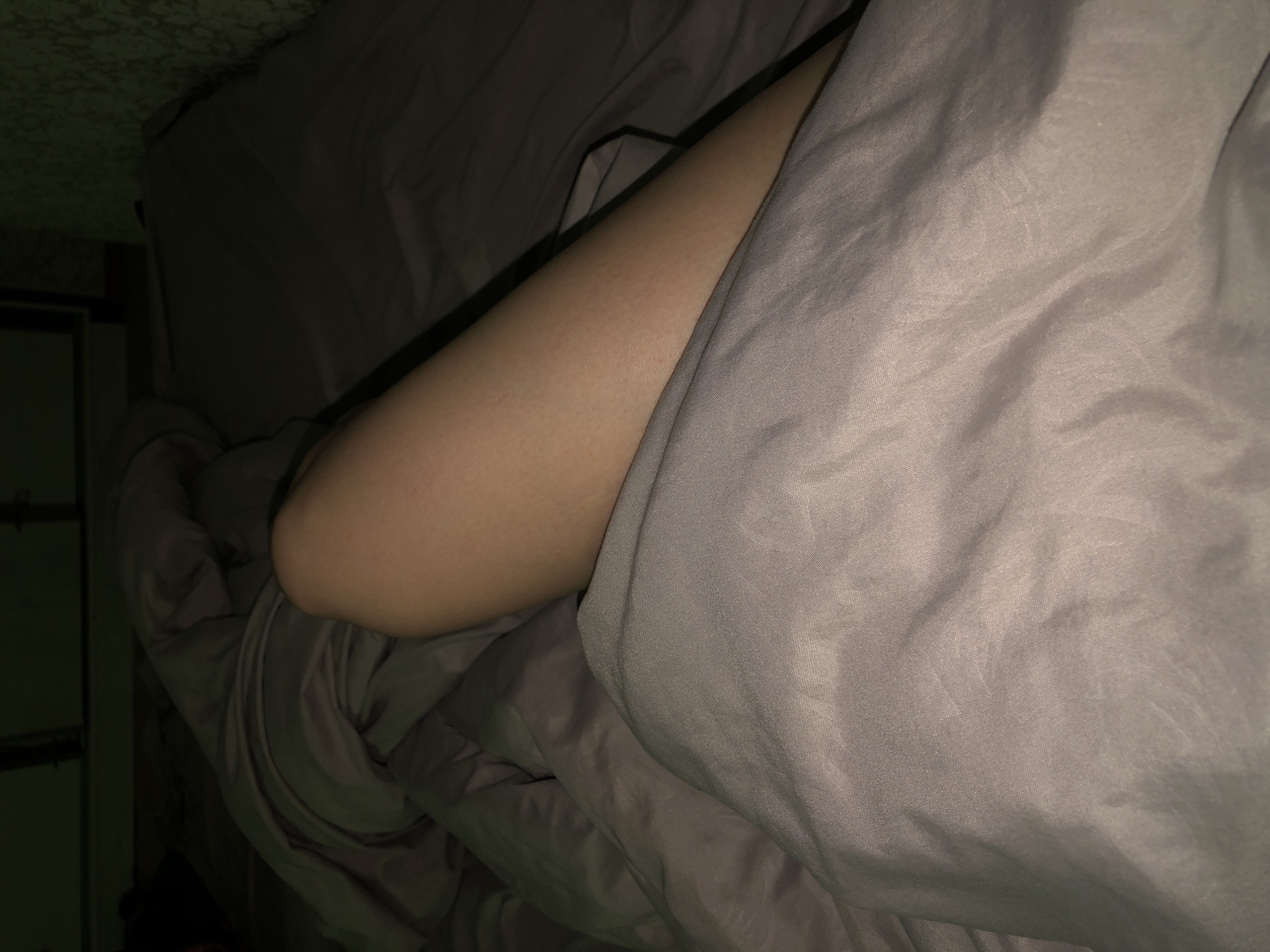 A human arm protrudes from beneath gray bedsheets. The scene is dimly lit. A cabinet is visible in the background. No further details are discernible about the location or city of Noviy, Russia.