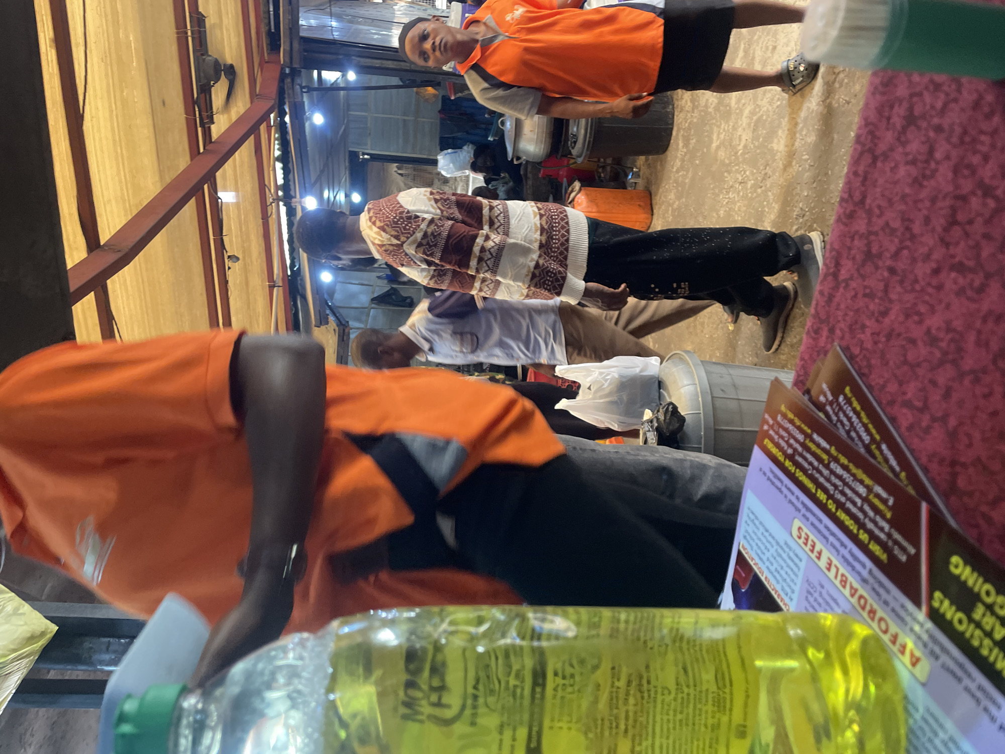 The image depicts a scene in an outdoor market or roadside stall, possibly in Abuja, Nigeria. In the foreground, a large plastic bottle filled with a bright yellow liquid, likely a cleaning product or beverage, is prominently featured. To the left, partially visible, is a person wearing an orange collared shirt. In the background, several individuals are present. One man wears a patterned sweater and dark pants. Another man is in a light blue shirt and beige pants. A woman in an orange and gray shirt stands on the right side of the frame, looking towards the camera. Various market elements are visible, including stalls, roofing structures, cooking pots, and plastic bags. The ground appears to be concrete or dirt. The overall atmosphere suggests a bustling, everyday commercial environment.