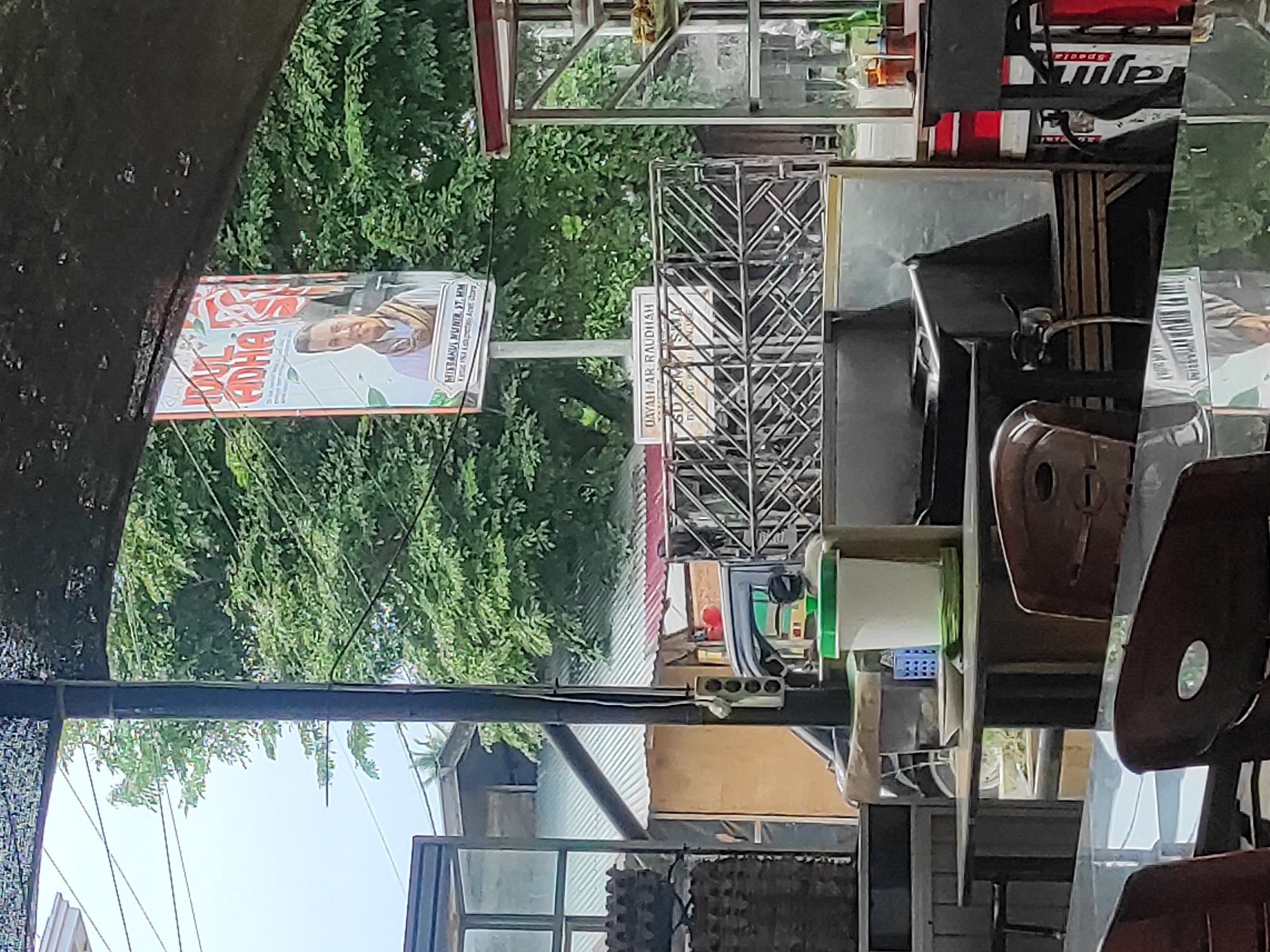 The image captures an outdoor scene likely within the city of Lhokseumawe, Indonesia, possibly near a food establishment. In the background, lush green trees dominate the scene, suggesting a tropical environment. A large banner is prominently displayed, featuring a portrait of a man wearing a headscarf and text in a foreign script, possibly related to a local event or candidate. Below the banner, a metal fence or railing is visible, separating different areas. Further in the foreground, tables and chairs suggest a seating area for patrons. Several objects are scattered on a table, including a white container with a green lid, a plate, and a small blue container. A vehicle is partially visible in the mid-ground. The overall impression is one of a casual, possibly bustling, outdoor public space.