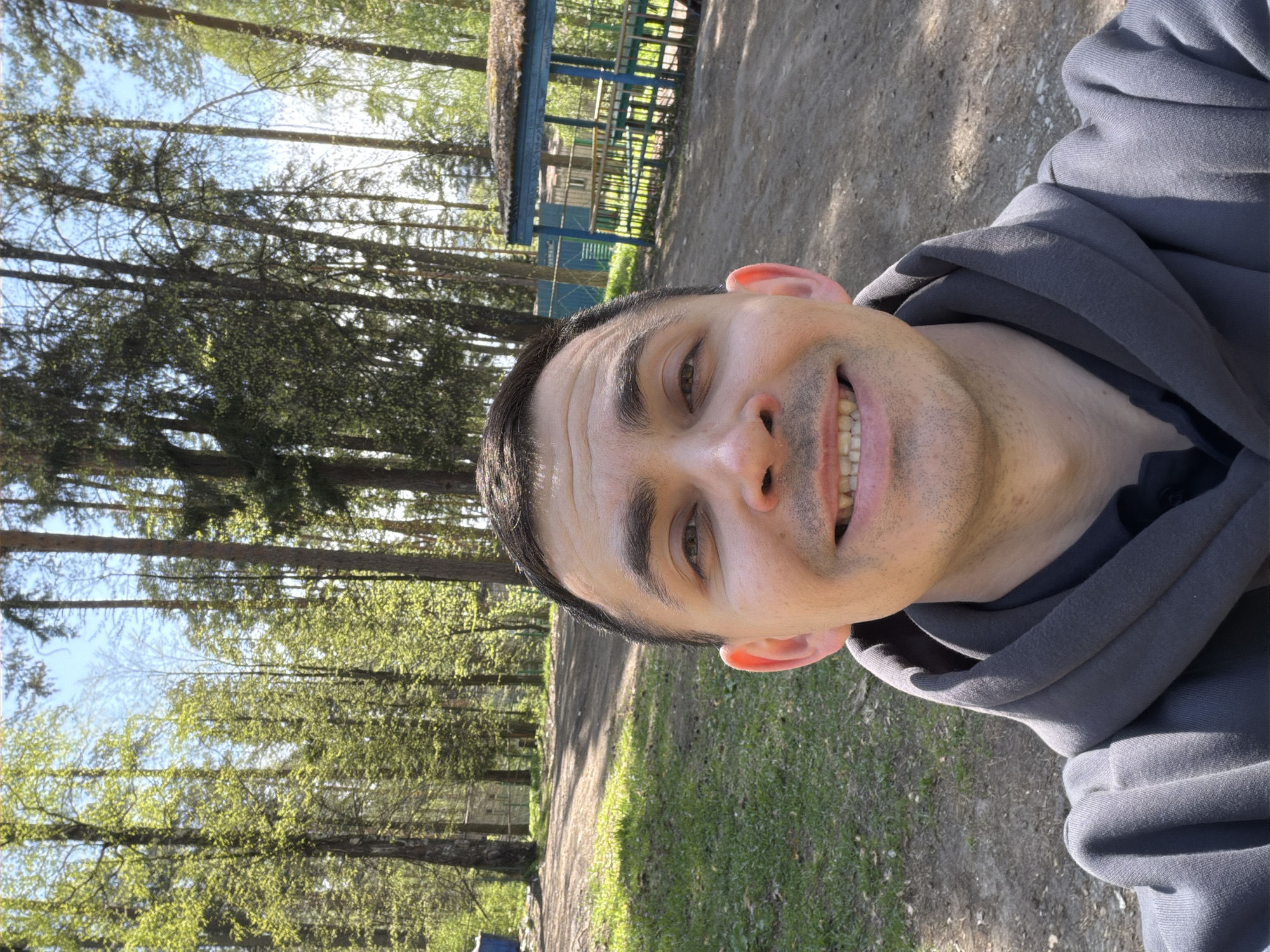 A man in a gray hooded sweatshirt smiles directly at the camera, with his head turned slightly to the right. Behind him is a forest with tall trees, some in full leaf. A dirt path leads into the trees. In the background, a blue structure with a sloped roof is visible. The location appears to be a park or wooded area in Izhevsk, Russia.
