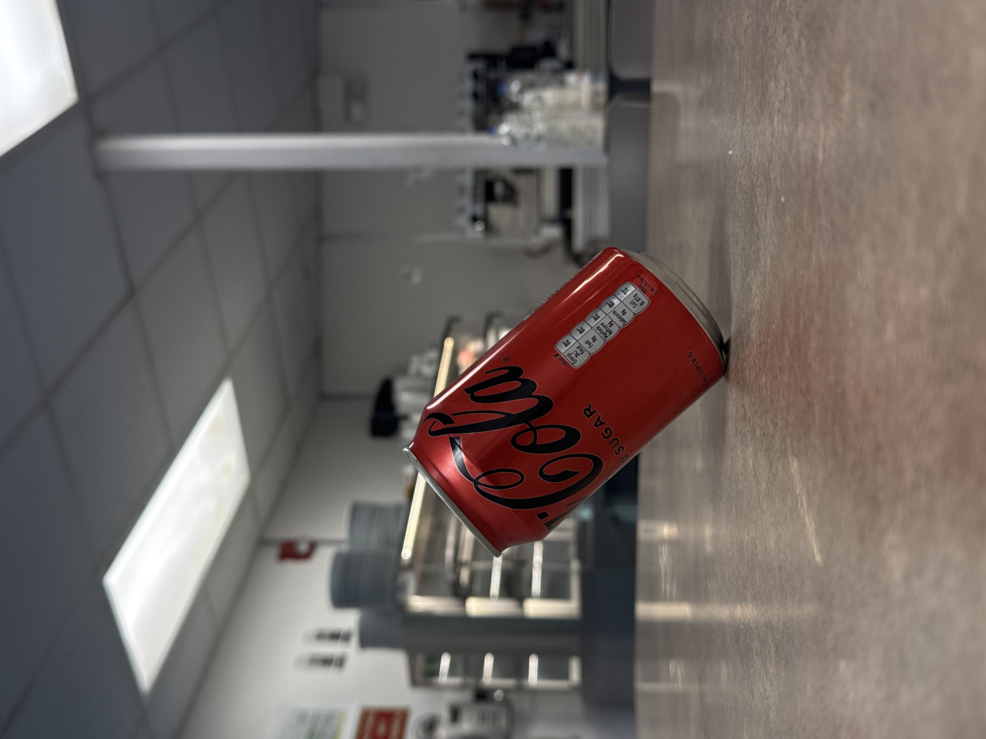 A close-up, eye-level shot shows a red can of Coca-Cola Zero Sugar resting at an angle on a speckled, light brown countertop. The can is positioned slightly off-center to the right. The background, out of focus, suggests an indoor commercial setting, possibly a cafe or a break room. Above the can, a rectangular fluorescent light fixture illuminates the ceiling, which is composed of square tiles. To the left of the can, stacked grey plates are visible, and further back, stainless steel shelving units and equipment are present, hinting at a food service area. A white column stands in the mid-ground, and beyond that, more shelving and bottles of water are faintly discernible. The overall lighting is bright, creating reflections on the countertop and highlighting the can. The environment appears clean and functional.