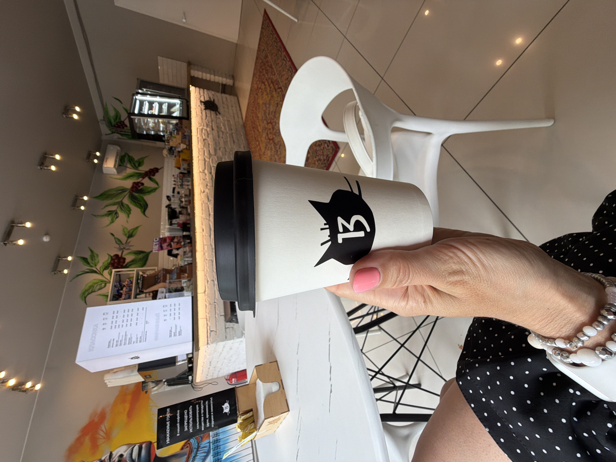 A close-up shot captures a hand holding a white coffee cup with a black lid and a black cat logo with the number "13" on it. The background reveals a modern cafe interior. To the left, a white table holds a wooden dispenser filled with sugar packets. Behind the table, a tall white sign displays text and a menu. The cafe wall features vibrant murals of coffee branches with red berries and a face. A white brick counter with another black cat logo is visible, and a refrigerated display case with desserts stands behind it. A red oriental rug lies on the tiled floor, and white chairs are scattered throughout the space. The image is shot in Birobidzhan, Russia.