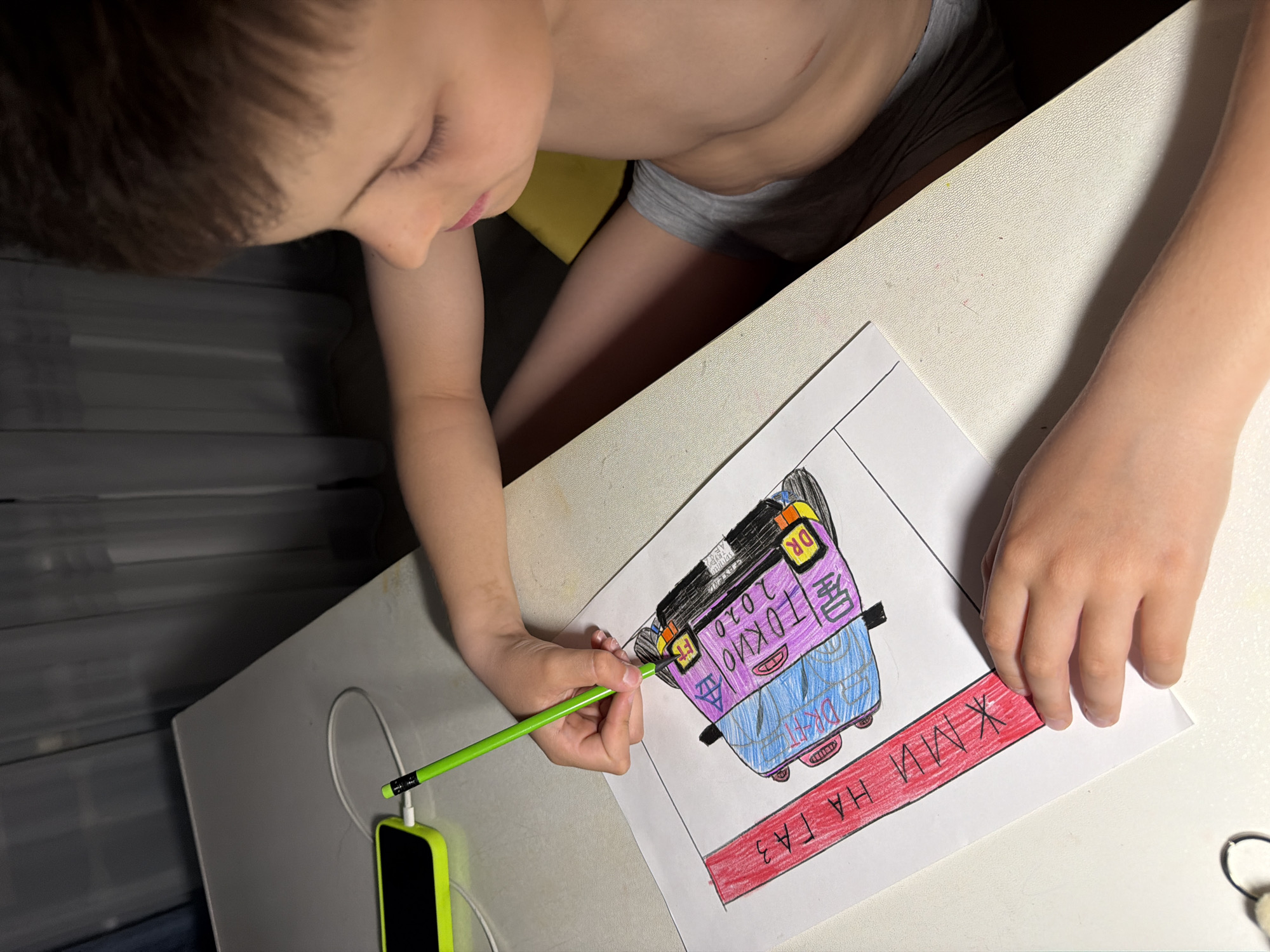 A child, viewed from above, is drawing on a sheet of paper on a white table. The child is fair-skinned with brown hair, wearing grey underwear. The drawing depicts a car with colorful markings and text. A green pencil rests on the drawing, and a yellow smartphone is connected to the pencil via a cable on the left side of the table. The location is unknown.