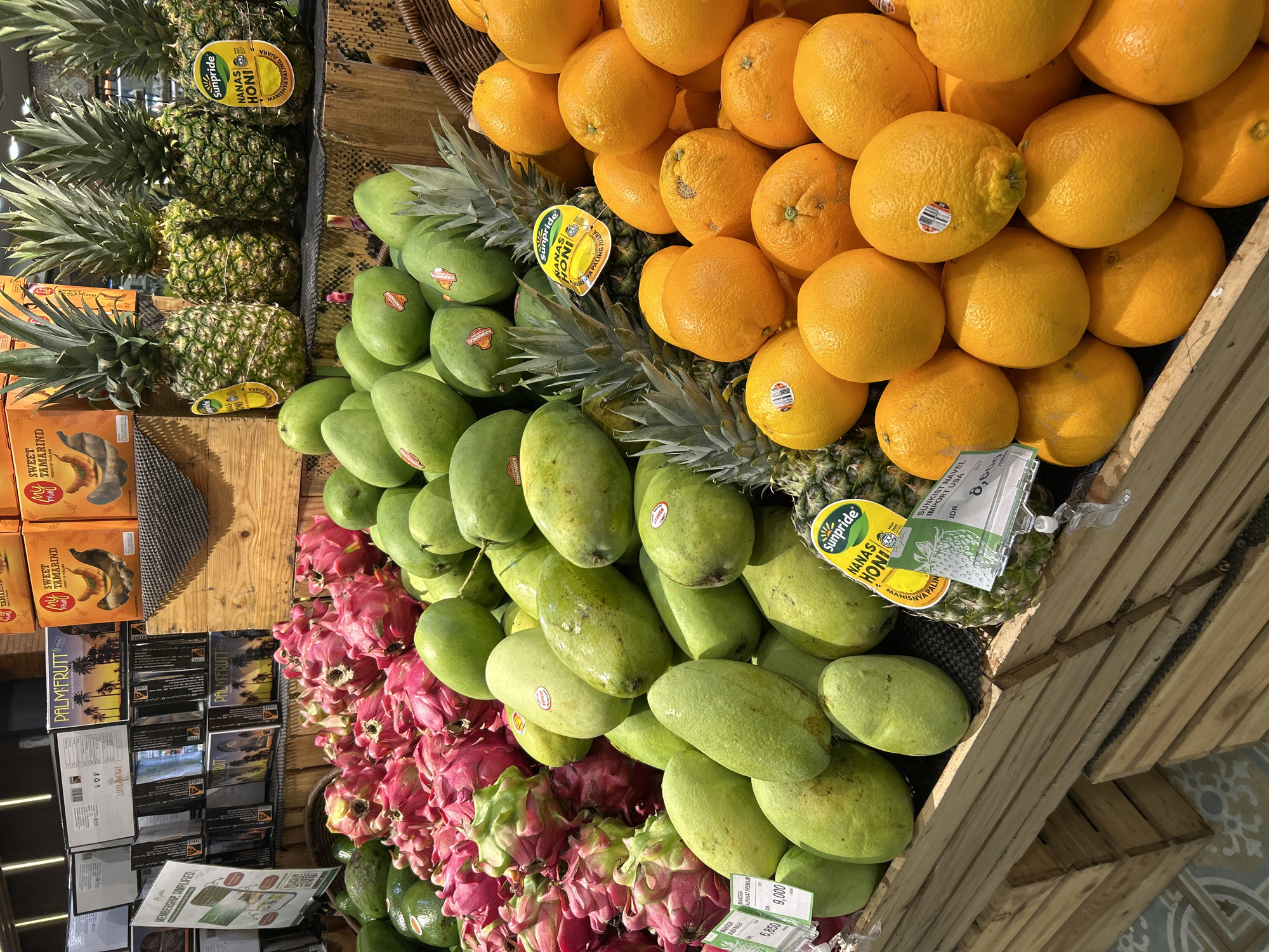 The image shows a vibrant display of fresh fruit at a market stall in the city of Ubud District, Indonesia. In the foreground, a pile of bright orange oranges is arranged in a wooden crate. Behind the oranges, a section of green mangoes is visible, some with stickers indicating their origin. Further back, pink dragon fruits are piled high, interspersed with more green mangoes. Several ripe pineapples, some with "Sunpride" stickers, are also part of the display. In the background, shelves are stocked with various boxed products, including what appear to be palm fruit and sweet tamarind. The overall impression is a colorful and abundant fruit market.