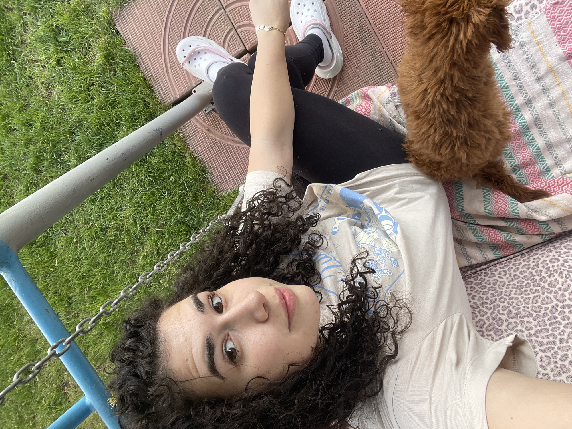 A high-angle, selfie-style photograph shows a woman with curly dark hair lying on what appears to be a swing or cushioned seat. She is wearing a light-colored t-shirt, black leggings, and white Crocs with pink accents. A small brown dog stands on the seat next to her. She has a bracelet on her right wrist. The swing is made of metal painted in blue and silver with hanging chains. In the background, there is green grass and a textured brown surface, likely the ground under the swing set. A patterned blanket is on the seat. The location is not evident in the picture. This image does not permit the identification of the city of Sofia.