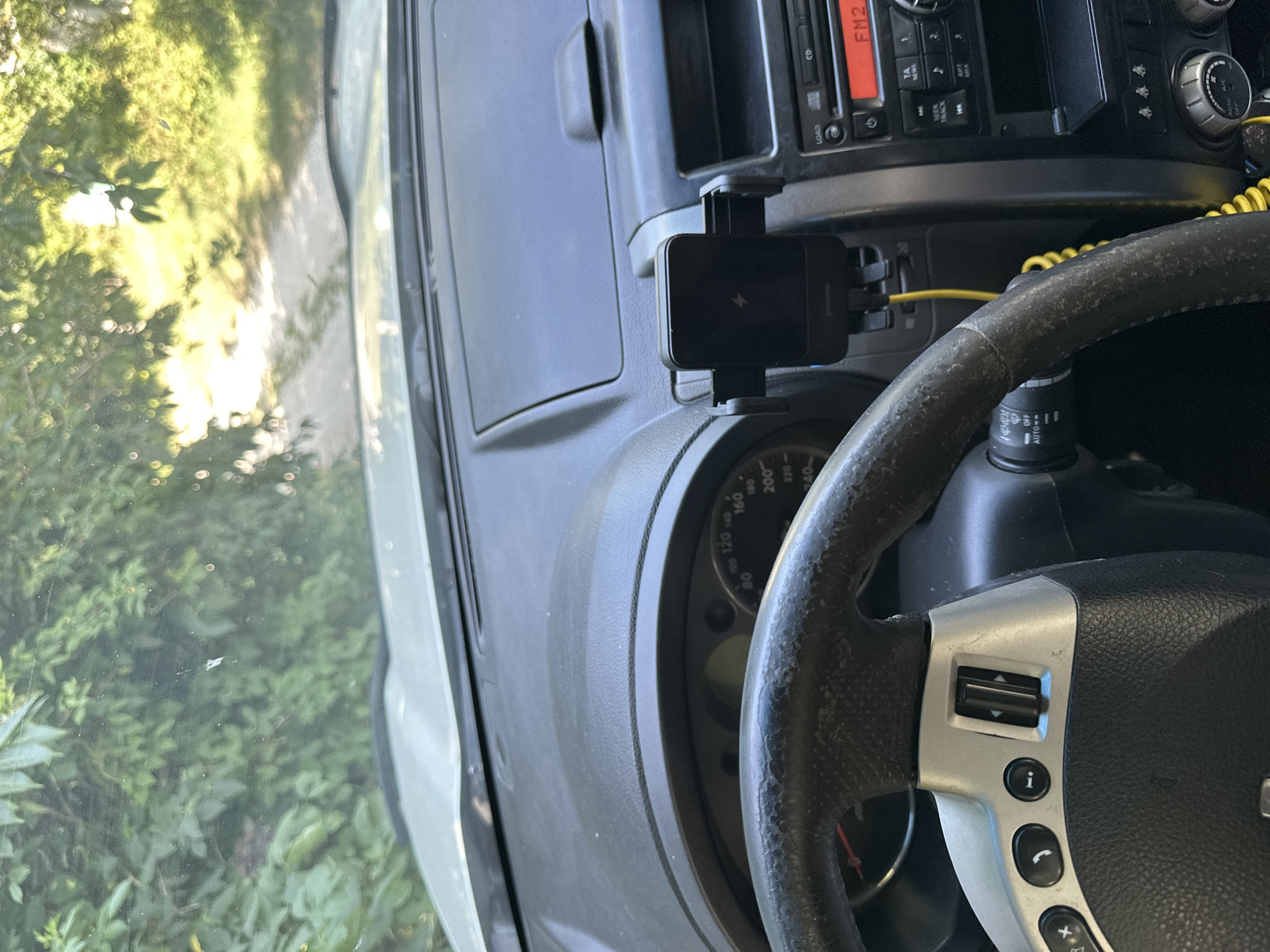 The image provides a view from inside a car, looking forward through the windshield. The car's dashboard is visible, featuring a steering wheel, instrument cluster, and a car radio. A black phone mount with a charging phone is attached to the dashboard. Outside the car, a sunlit dirt road is visible, lined with trees and foliage. The scene appears to be in a rural area. The specific location within Makiv, Ukraine is not discernible from the image alone.
