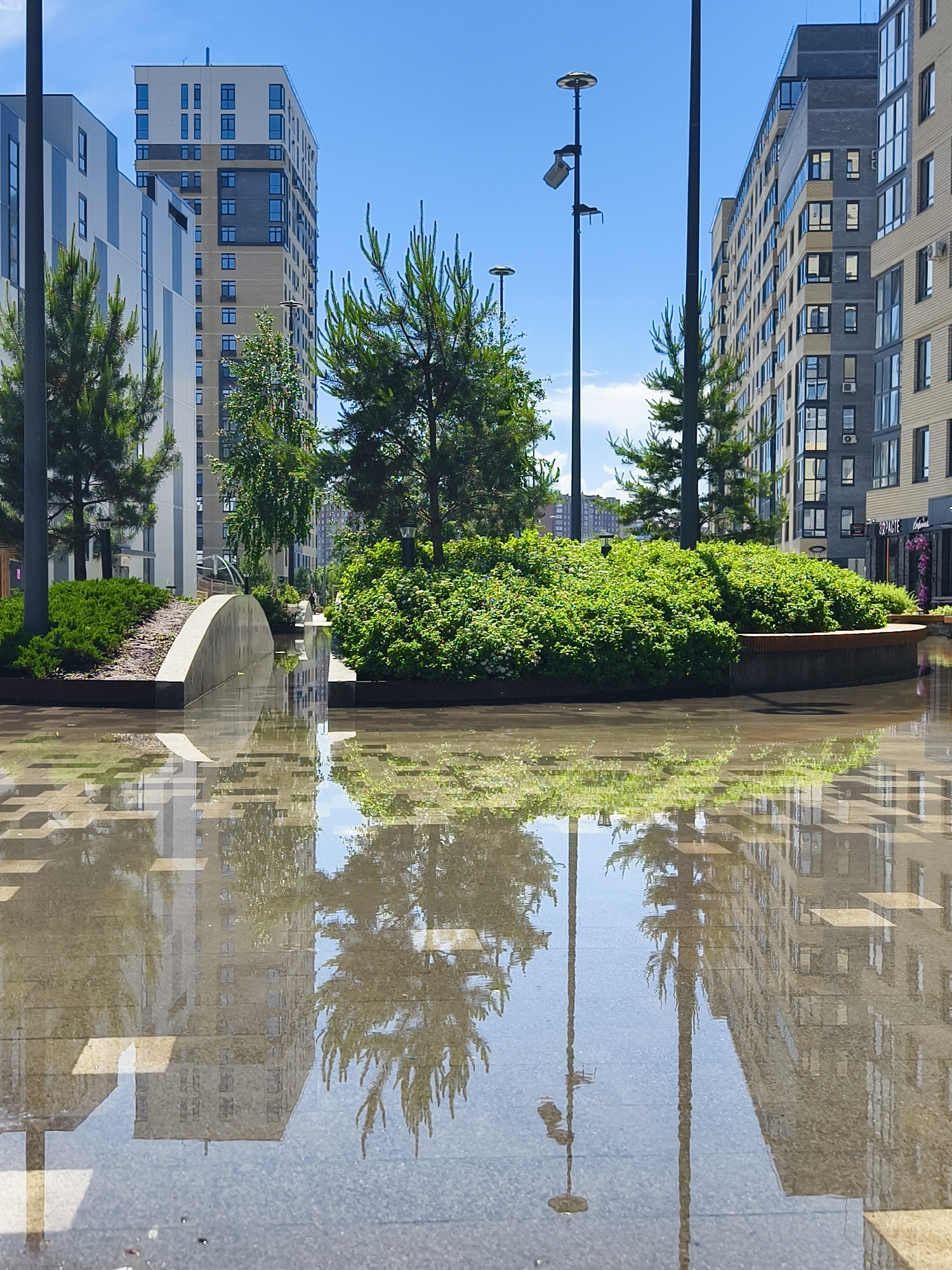 This vibrant vertical shot captures a modern urban landscape in Tyumen, Russia, likely within a residential complex or public park, under a bright blue sky. The scene features contemporary multi-story apartment buildings with varied facade textures and window designs, alongside abundant greenery including tall pine trees, deciduous trees, and manicured bushes arranged in landscaped beds.
The foreground is dominated by a large, smooth surface of water, resembling a puddle or a shallow water feature, which creates a striking mirrored reflection of the entire upper scene. This reflection perfectly duplicates the buildings, trees, and streetlights, creating an abstract and symmetrical composition. One prominent feature is a low, curved concrete barrier or planter wall dividing two sections of green foliage near the center of the image. Tall, slender streetlights with multiple fixtures are visible, both in the actual scene and their reflections.
No people or activities are explicitly taking place, lending a calm and serene atmosphere to the scene. The bright sunlight suggests it is daytime, likely mid-morning or early afternoon, with clear weather despite the wet ground. On the rightmost building, a commercial sign is visible with Cyrillic text that appears to read "БИРЖА СТЕЙКОВ" (Steak Exchange) and below it, "РЕСТОРАН" (Restaurant), indicating a business establishment. The overall impression is one of modern urban planning with a focus on green spaces and aesthetic design.