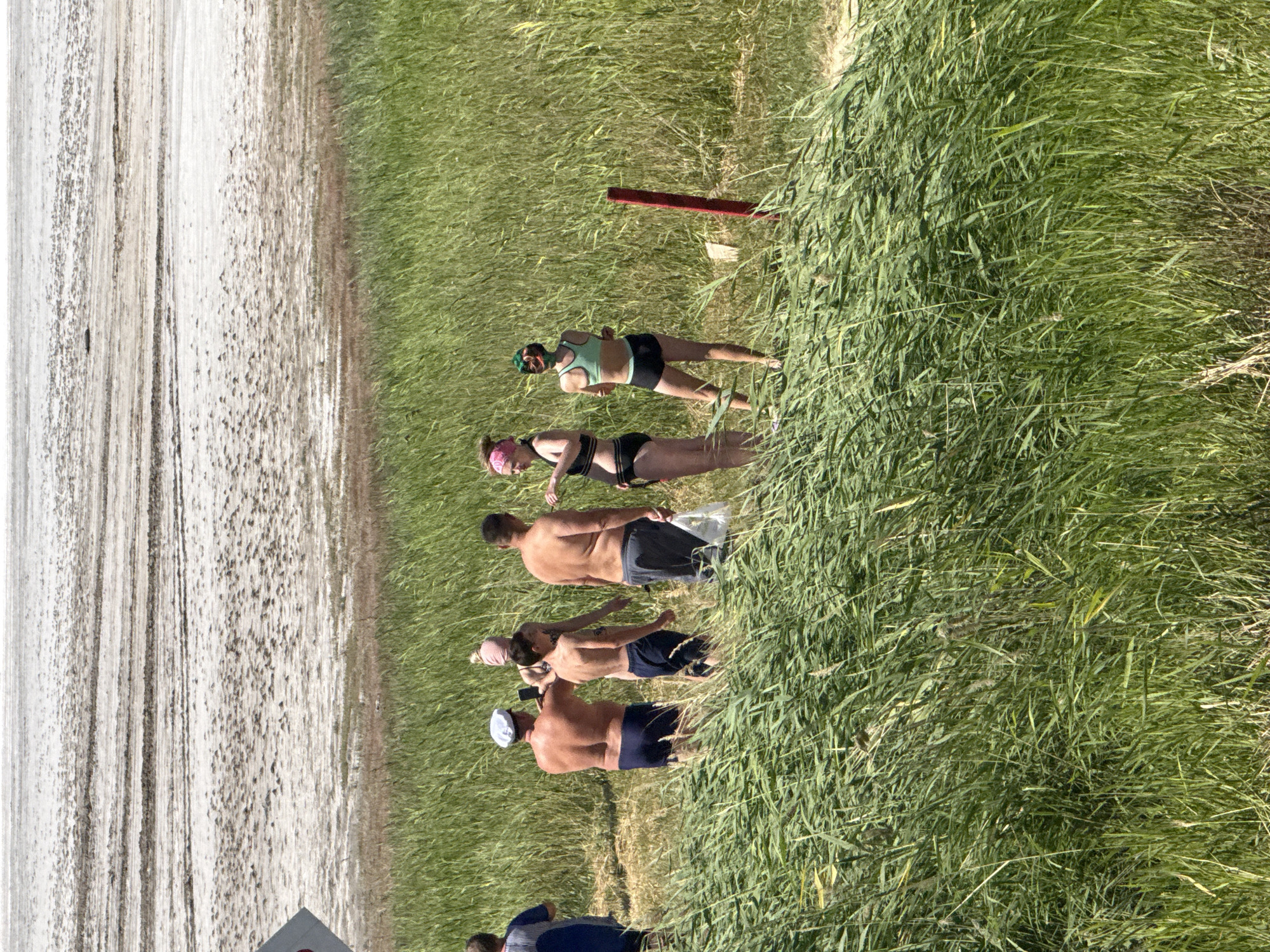 In Бурсоль, Russia, a group of people are gathered on a beach near a body of water with a light-colored, textured ground. The foreground is dominated by tall, green reeds and grass. From left to right, a person in a blue shirt is visible. Next, a man wearing a white hat and blue swim trunks stands with his back to the camera, holding a phone as if taking a picture. Beside him is a shirtless boy wearing blue shorts. To their right, a shirtless man in grey shorts stands with a white plastic bag in his hand. Further to the right, two women are positioned. One woman wears a black bikini and has a pink headband. The other woman is in a green swimsuit top and black shorts, with a colorful head covering. A red post is visible in the grass behind the women. The background shows a vast, flat expanse of textured, light-colored ground, likely salt flats or mudflats, stretching towards a hazy horizon.