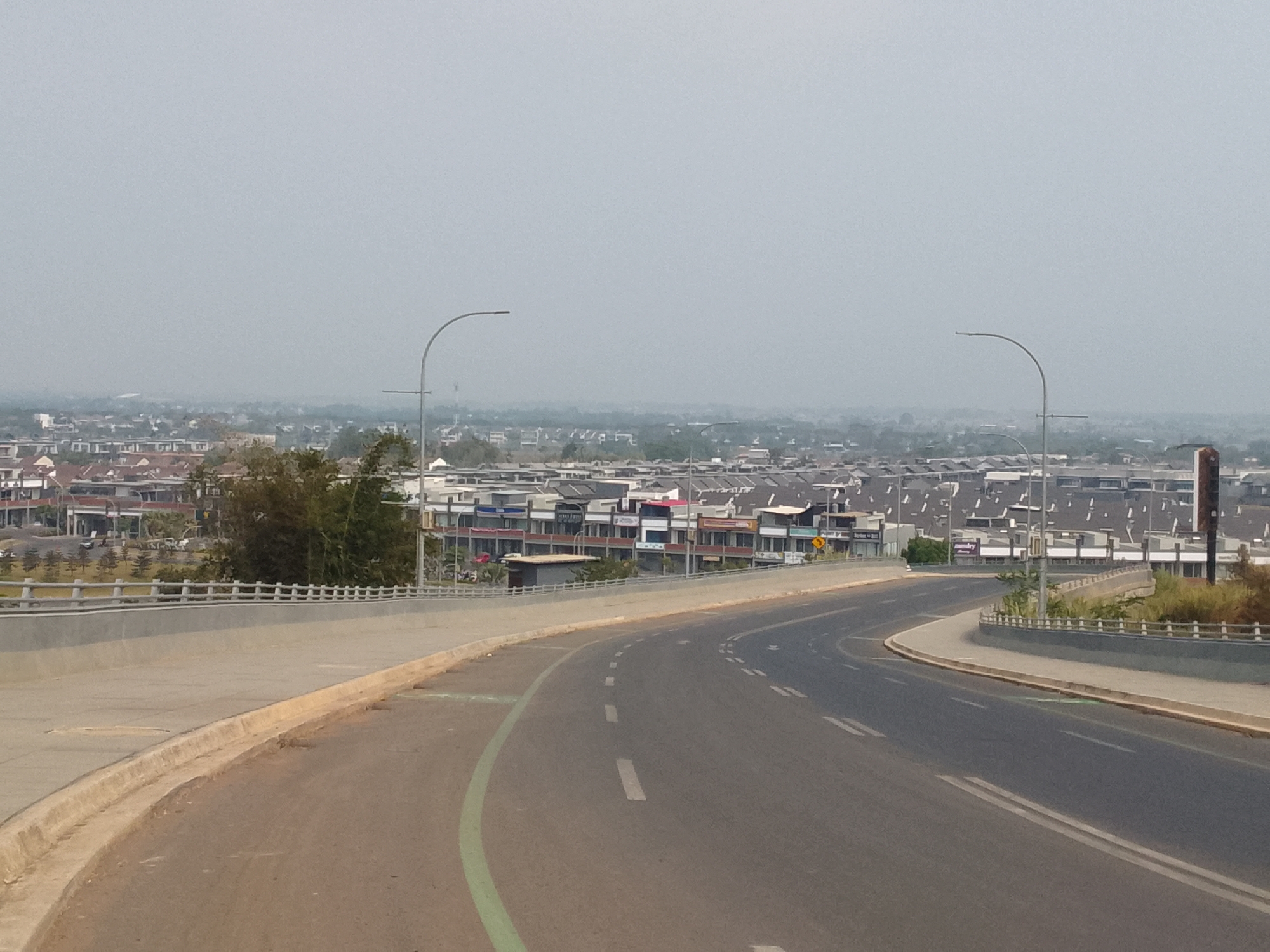 The image captures a view of a road leading into a developed area in Bojonghaleuang, Indonesia. The road curves to the right, with a concrete barrier and a sidewalk on the left. Streetlights are visible on either side of the road. In the background, a dense collection of multi-story buildings, likely residential and commercial properties, stretches across the landscape under a hazy sky. Some of the buildings appear to be shops or small businesses. There are no people or vehicles clearly visible in the frame. The environment appears to be a developed urban or suburban area.