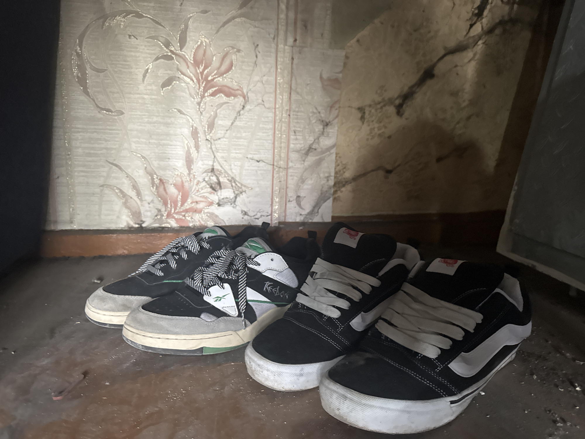 The image depicts a pair of Reebok and a pair of Vans shoes resting on a dusty, dark surface. The shoes are positioned close together, almost touching. The background consists of aged walls with floral wallpaper on the left and cracked, peeling paint on the right. The setting appears to be an enclosed space, possibly a neglected corner or storage area. Location is unknown.