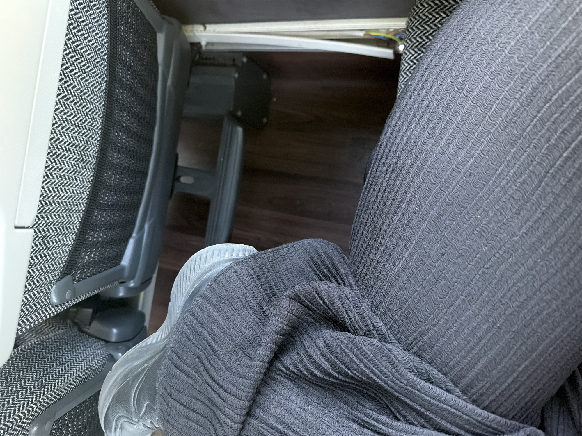 This image shows a close-up of a person's legs and feet resting in what appears to be an airplane or train seat. The person is wearing dark grey, ribbed pants and light grey shoes. The seat fabric is a dark grey and white herringbone pattern. Underneath the seat, some mechanical components and wiring are visible, along with a wooden floor. The scene does not provide any information to suggest it is located in Shtarkovo, Bulgaria.