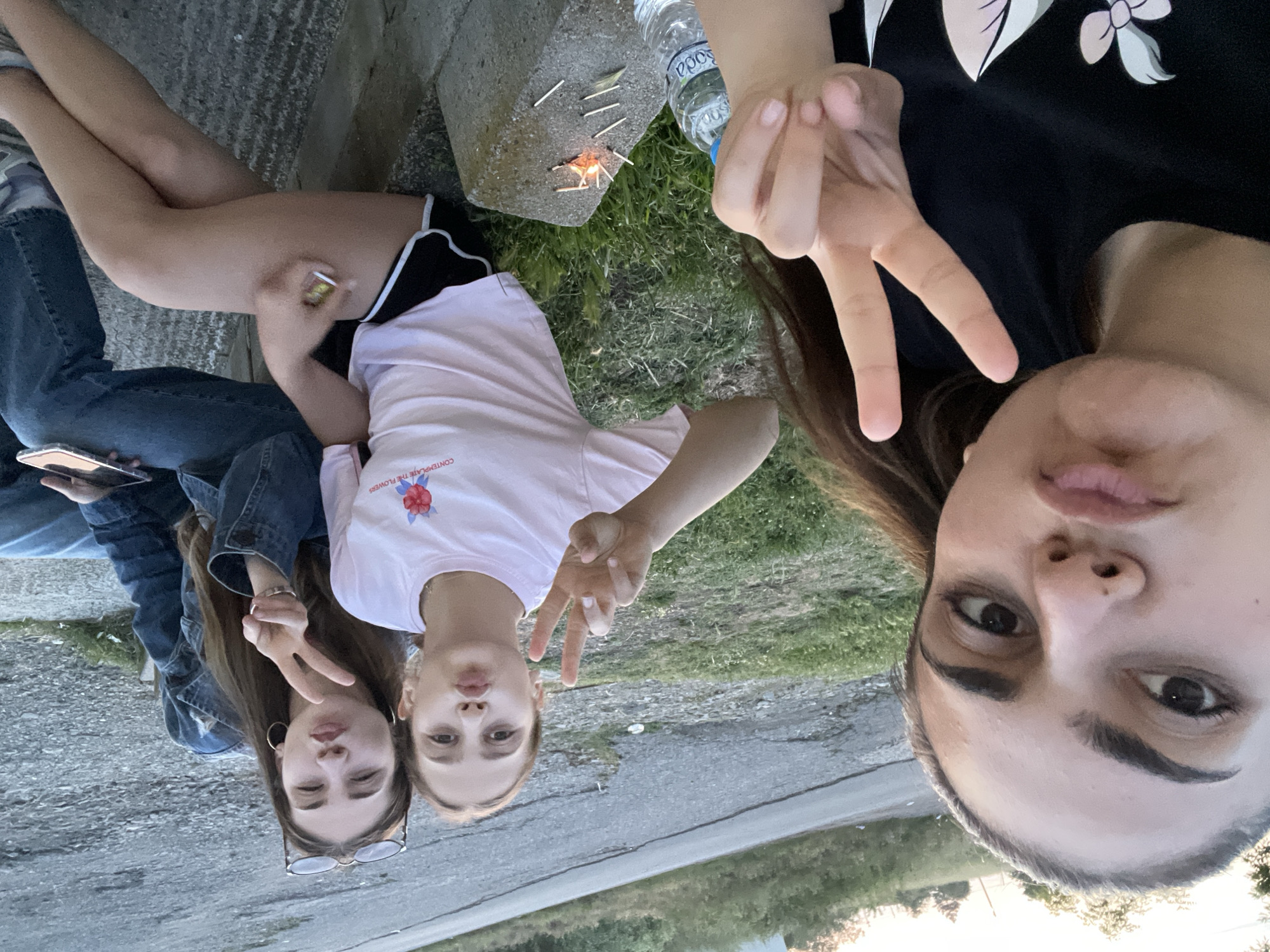 Three young females are captured in a selfie-style photograph taken outdoors in Debelt, Bulgaria. The subject closest to the camera, positioned on the left, has dark hair, wears a black t-shirt, and holds her right hand in a peace sign while pouting. To her right, a second female with light brown hair wears a light pink t-shirt featuring a red flower graphic and the text "CONTEMPLATE THE FLOWERS," along with black shorts with white trim. She also pouts and displays a peace sign with her left hand. She is seated on a concrete surface or curb, next to a grey concrete block on which several matches are scattered, some appearing lit with a small flame or glowing embers. A plastic water bottle, branded "Soda," rests near the concrete block. The third female, on the far right, has long brown hair, wears a denim jacket, blue jeans, and large hoop earrings. Sunglasses are perched on top of her head. She mirrors the others with a pouting expression and a peace sign from her right hand, while holding a light-colored smartphone in her left hand. All three subjects are looking towards the camera. The background features an asphalt road, grassy embankments, and distant trees, illuminated by what appears to be late afternoon or early evening light.