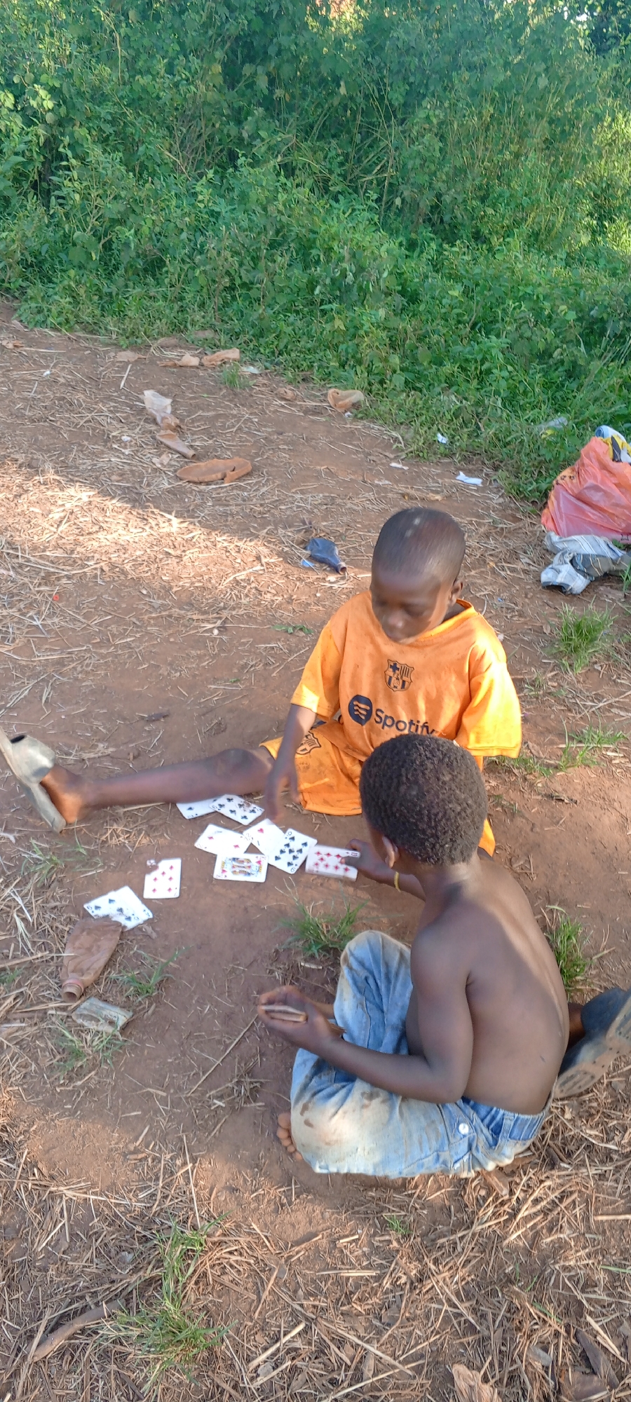 Two children are playing cards on bare earth. One child wears an orange t-shirt with the FC Barcelona and Spotify logos. The other child is shirtless and wears blue jeans. The ground is littered with debris and dry organic material. In the background is dense green vegetation. The location is in Ngalula, Democratic Republic of the Congo.