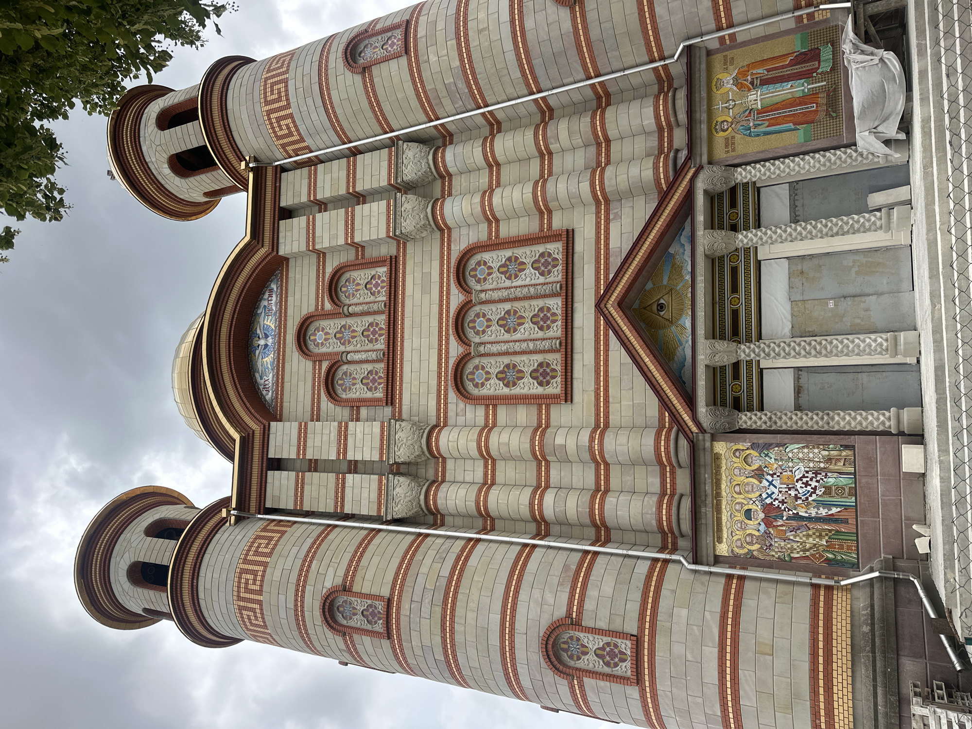 The image shows the facade of the Church of the Nativity of the Blessed Virgin Mary in Ternopil, Ukraine. The church is a large, ornate building with two towers and a golden dome. The facade is decorated with mosaics and colorful tiles. The photograph appears to be taken from a low angle, looking up at the church. The sky is overcast. Construction materials are visible in the foreground, suggesting that the church is either under construction or undergoing renovations.
