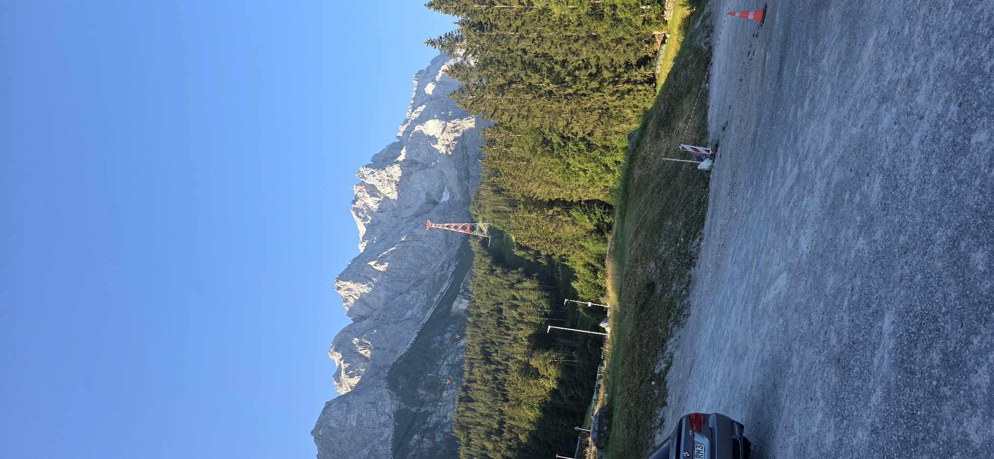 In Grainau, Germany, a panoramic view showcases the majestic Zugspitze mountain range under a clear blue sky. A prominent red and white cable car pylon stands tall on the mountainside, amidst a dense forest of evergreen trees. In the foreground, a gravel parking lot is visible, with a dark-colored car parked on the left side, its license plate reading "RM3". Several traffic cones and a barricade are placed on the edge of the parking lot, indicating ongoing work or a restricted area. The sun casts long shadows, suggesting it is either early morning or late afternoon.