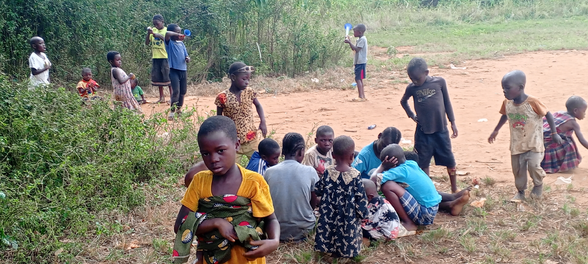 A group of children is gathered in a clearing in Ngalula, Democratic Republic of the Congo. The ground is dirt and sparse vegetation. In the foreground, a child with a yellow shirt is holding a bundle wrapped in patterned fabric. Behind them, a group of children are sitting on the ground, and others stand around them. Some are barefoot. To the left, there are dense bushes and to the right, the ground opens into a larger clearing. Several other children are further back, and some are holding up small objects.