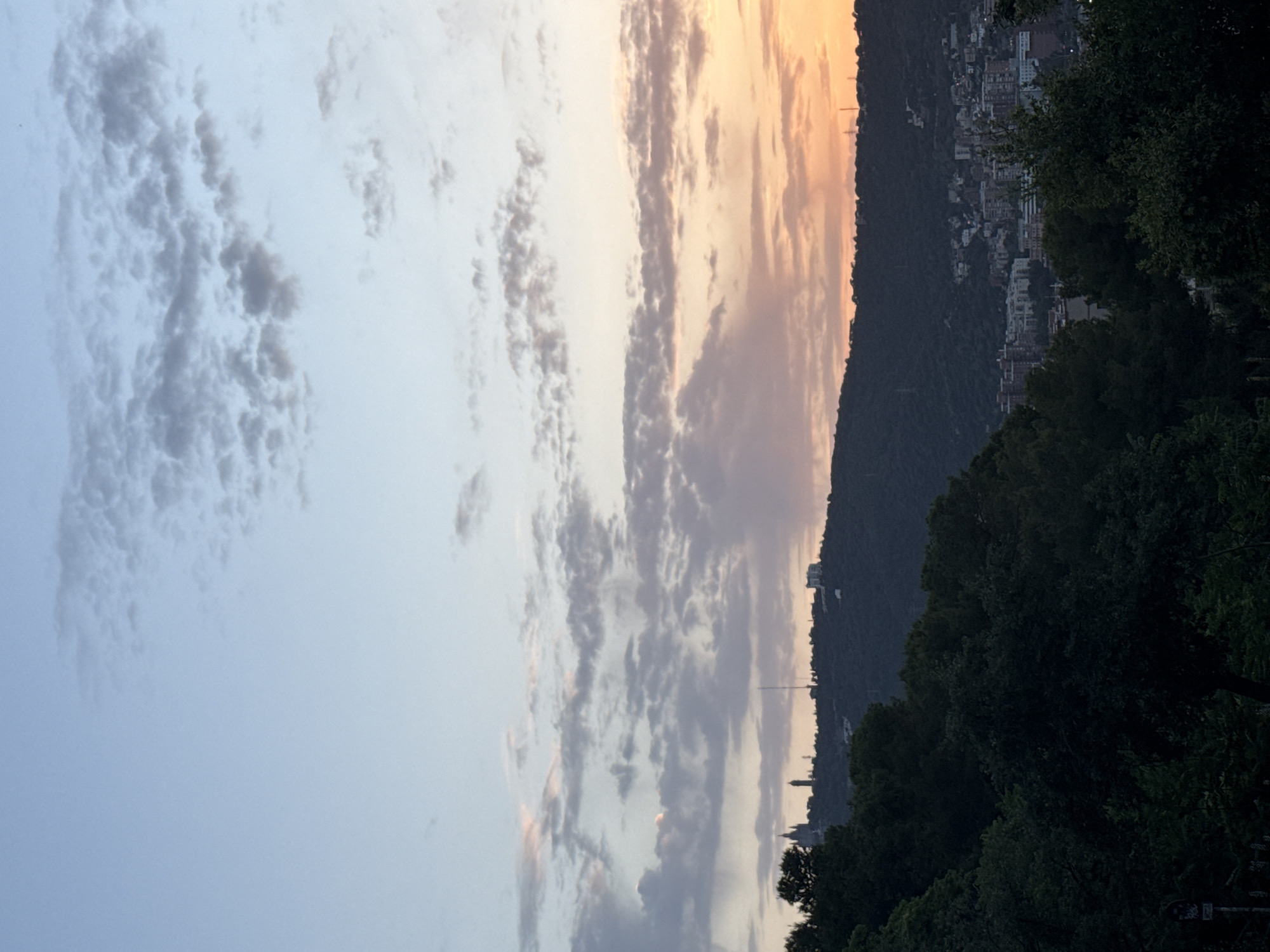 This image depicts a panoramic view of the city of Barcelona, Spain, likely taken during either sunrise or sunset given the warm hues in the sky. The foreground is dominated by dense, dark green trees, obscuring the lower portion of the city.
The middle ground features a prominent, tree-covered hill or mountain range that forms a dramatic backdrop. On the crest of this hill, several structures are visible. To the left, there is a large, imposing building with multiple towers, which resembles a religious or historical monument, possibly the Tibidabo Temple or a similar landmark. Further along the ridge, a tall antenna or telecommunications tower stands out. Beyond these, a cluster of buildings, indicative of a town or urban development, is spread across the hillside.
The sky above is a gradient of soft blues and pastel shades of orange and pink, with scattered clouds that are illuminated by the setting or rising sun. The overall impression is one of a serene and picturesque urban landscape.