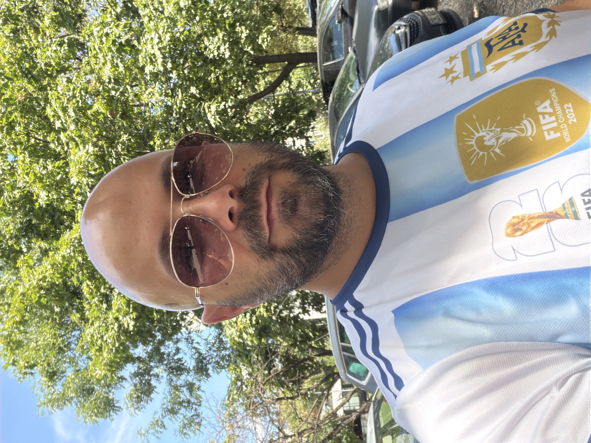 A bald, middle-aged man with a beard and sunglasses is pictured outdoors, wearing a white and light blue striped jersey with the FIFA World Cup 2022 logo and the Argentinian Football Association (AFA) crest. He is standing in front of trees and parked cars. The scene appears to be a street in Sofia, Bulgaria.