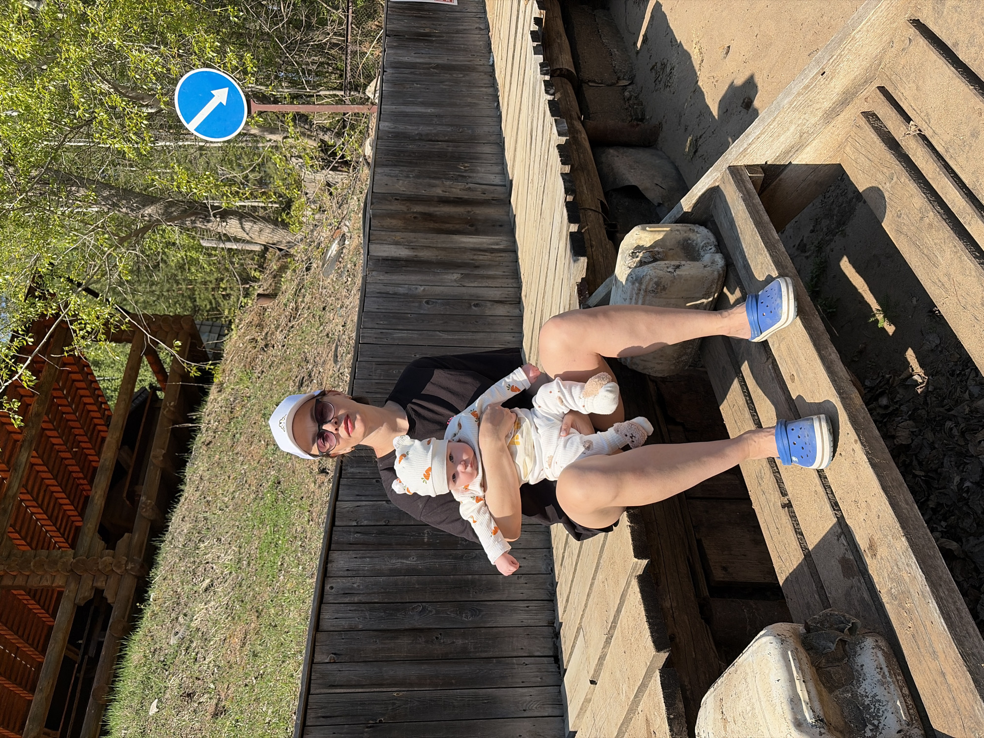 Outside in daylight, a fair-skinned adult wearing a black shirt, white baseball cap, and sunglasses is seated, holding a baby dressed in a white outfit with carrots printed on it, with a matching hat. The adult has bare legs and wears blue Crocs. They are on a wooden platform with a wooded area to the left and a road sign indicating direction to the top left. The location is Chaikovsky, Russia.