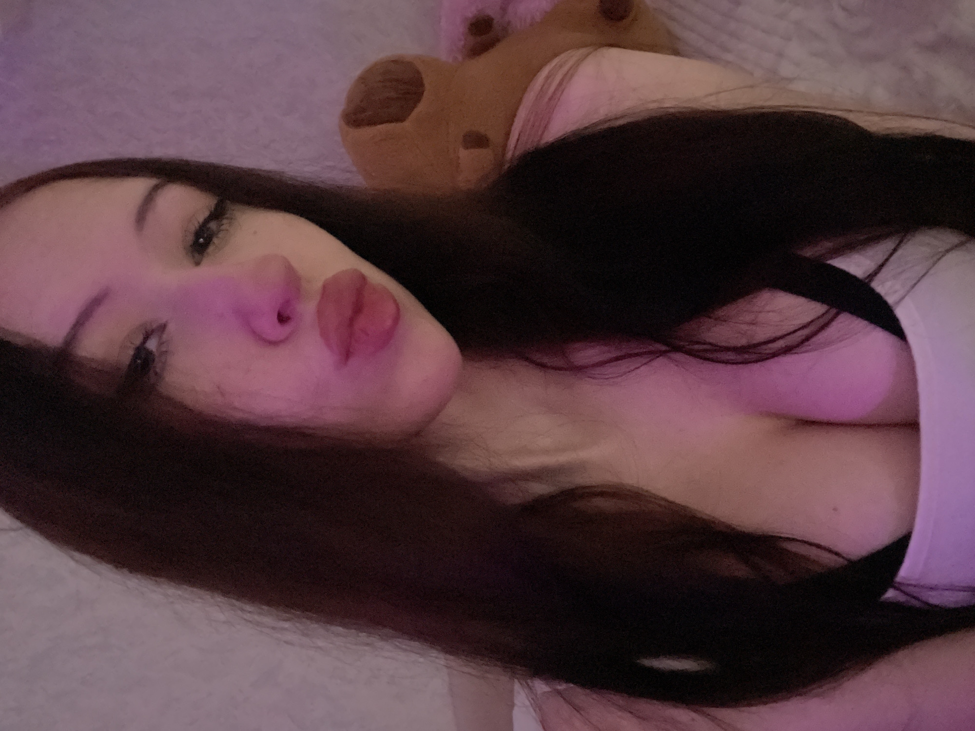 The image is a close-up selfie of a young woman with long, dark hair and fair skin. She is pouting her lips and looking towards the camera. She is wearing a white top and a black bra. In the background, there is a brown teddy bear and a pink blanket. The lighting is dim and appears to be from artificial sources, casting a pinkish hue on her face. The overall setting suggests an indoor environment, possibly a bedroom. There is no visible indication of the location being in Votkinsk, Russia.