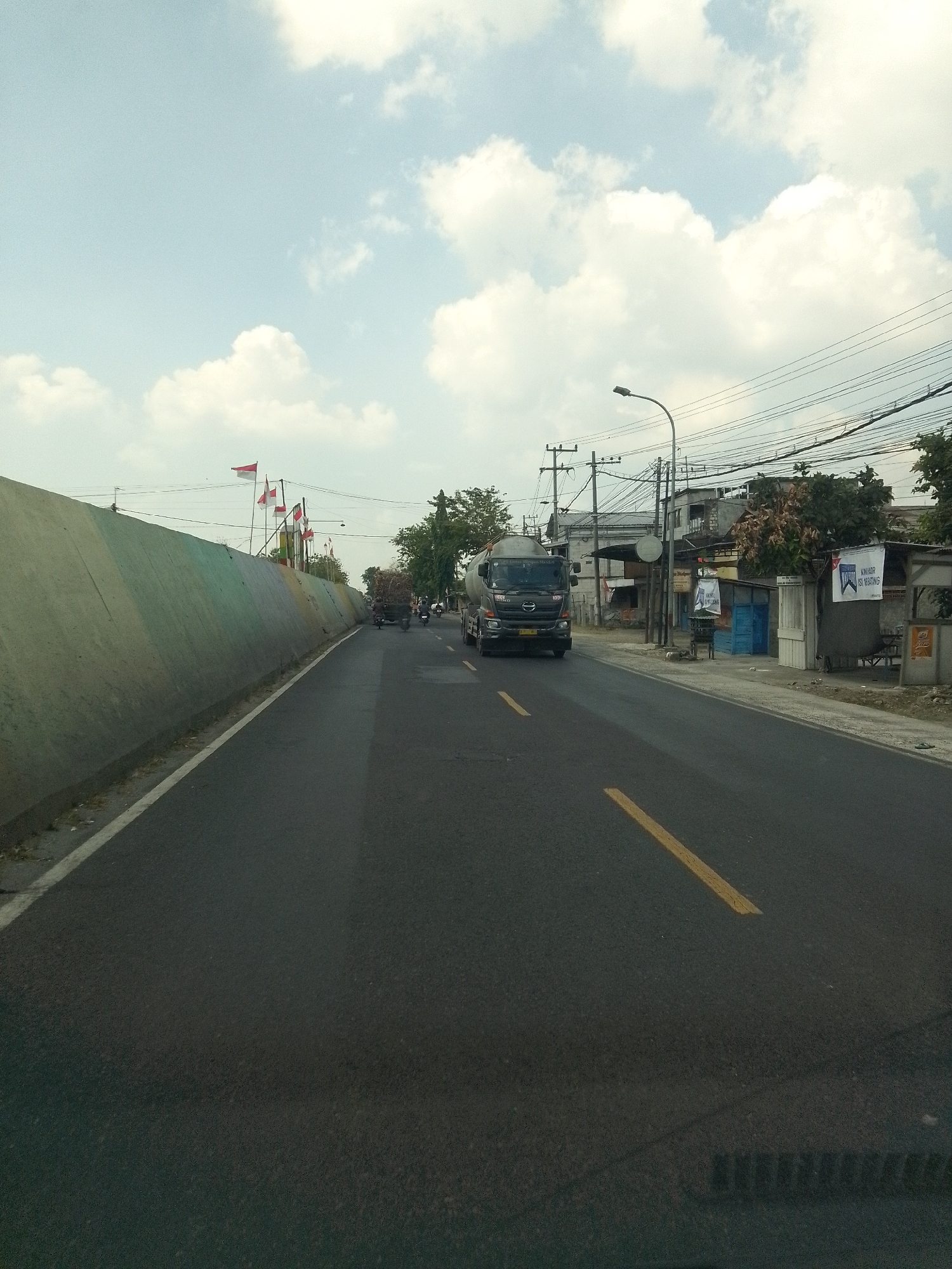 The image depicts a road in Banaran, Indonesia, under a bright, cloudy sky. A grey cement barrier runs along the left side of the road, which is paved with asphalt and marked with a yellow line. Several Indonesian flags are visible on poles atop the barrier. On the right side of the road, there are buildings, power lines, a lamppost, and trees. A cement truck is driving on the road, and in the background, another truck is visible. A motorcycle is also present on the road. The scene appears to be a view from a vehicle traveling along the road.