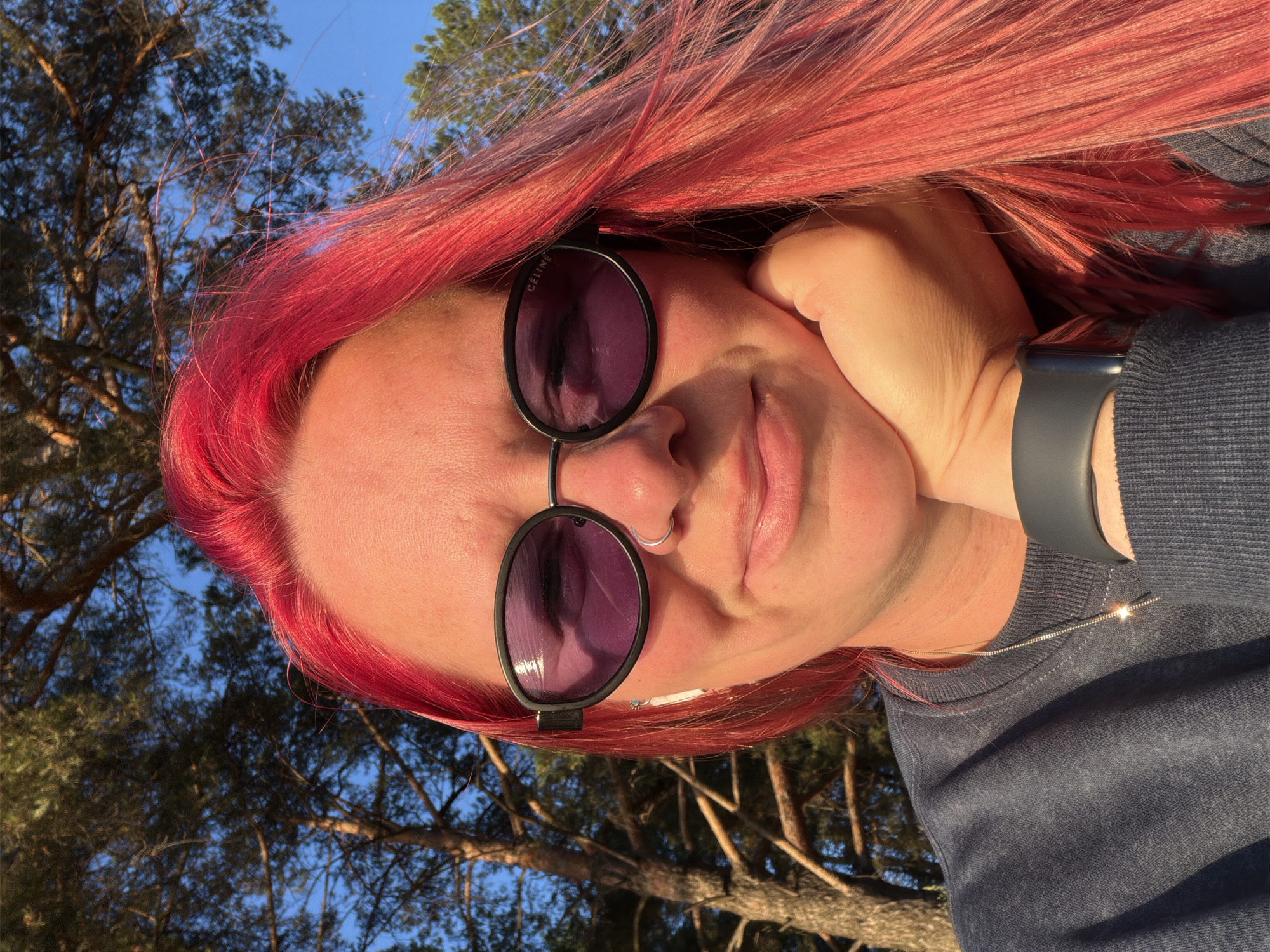 A close-up, outdoor, selfie-style photograph shows a person with bright red hair, wearing dark sunglasses with purple lenses, a nose ring, and a dark crewneck sweatshirt. The person's right hand is supporting their chin, and their left ear is visible with a white wireless earbud. The background consists of trees with sunlight filtering through their branches, and a clear blue sky. The lighting suggests it is either sunrise or sunset, casting a warm glow on the subject's skin. The overall scene is consistent with a natural environment, possibly a park or wooded area within the city of Chaikovsky, Russia, given the presence of pine trees.