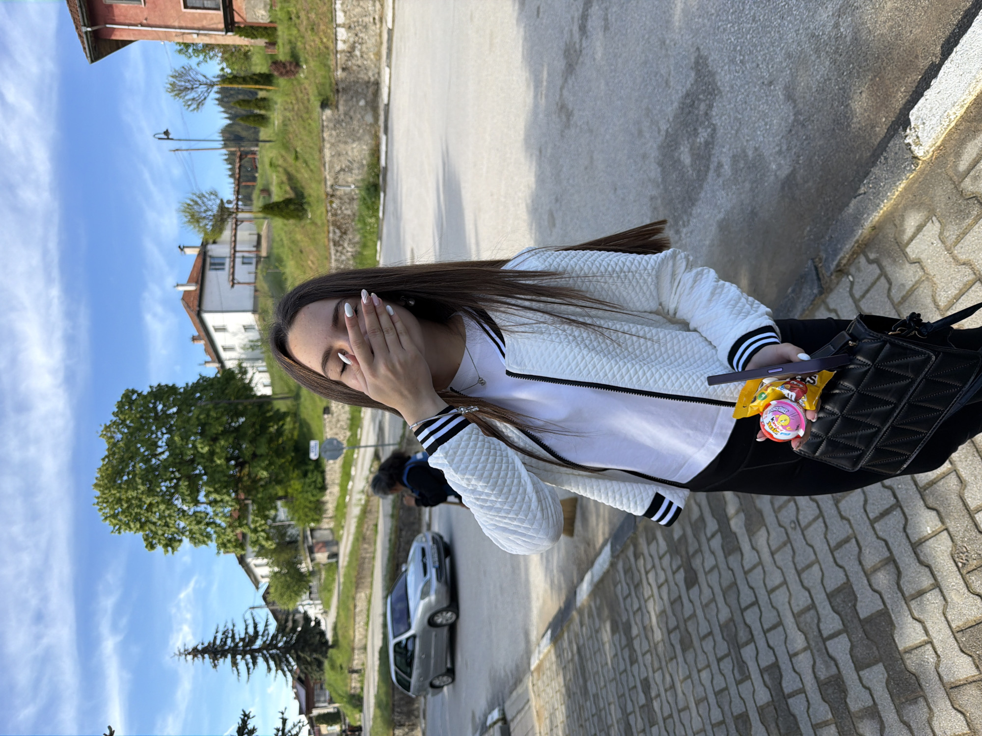 A young woman with long brown hair stands on a brick sidewalk in what appears to be Zmeitsa, Bulgaria. She covers her face with her right hand. She wears a white jacket with black and white striped cuffs over a white t-shirt and black pants. A black purse hangs from her shoulder. In her hand, resting on the purse, she holds a smartphone, a Kinder egg, and some wrapped candy. The background includes a road with a parked silver car, houses, trees, and a blue sky with light clouds.