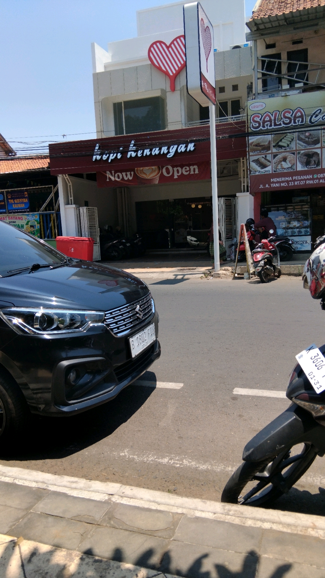 The image depicts the facade of a coffee shop named "Kopi Kenangan" in Batang, Indonesia. The shop features a red awning with the text "Now Open" and a stylized red heart logo above it. A black car is parked in front of the shop, partially obscuring the entrance. A motorcycle is also visible on the right side of the image, and another motorcycle is parked further inside the shop's premises. The street in front of the shop is paved and appears to be in an urban setting, with buildings and trees visible in the background. The sky is clear and blue, indicating a sunny day.