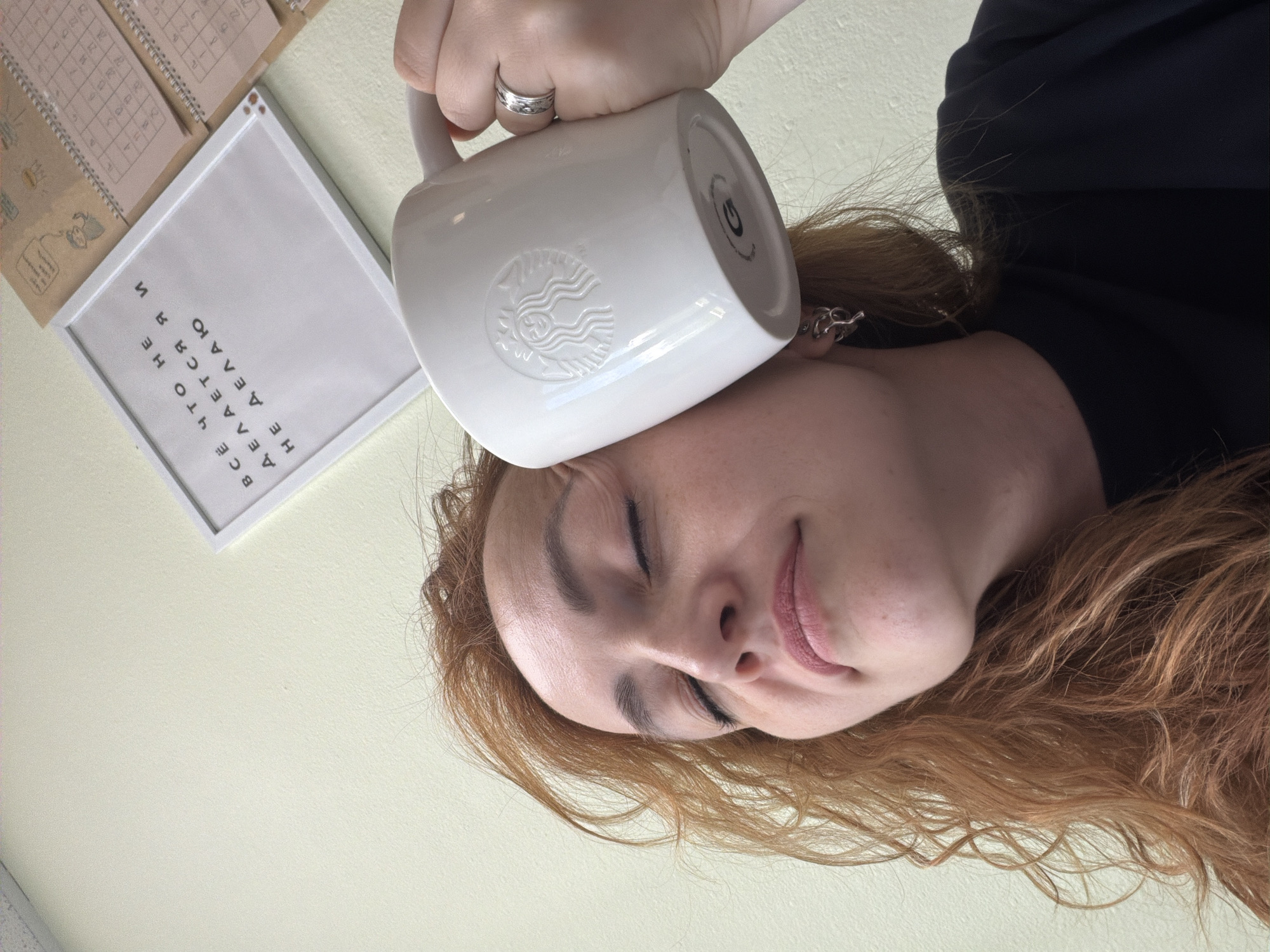 A young woman with reddish-brown hair is seen from a low-angle, upside-down perspective. She has her eyes closed and is smiling gently, with light pink lipstick on her lips. A white Starbucks mug is held in her right hand, positioned on top of her forehead. She is wearing a silver ring on her ring finger and a silver earring with a chain-like design in her left ear. The background is a pale, neutral color, likely a wall. To the left of the woman, there are two spiral-bound calendars or planners with grid-like pages, and a framed picture with text. The text in the frame, oriented to be read correctly from the viewer's perspective, says "ВСЁ ЧТО НЕ ДЕЛАЕТСЯ — К ЛУЧШЕМУ" which translates to "Everything that is not done is for the best." The overall scene suggests a casual, indoor setting. The lighting appears to be soft, possibly from natural light, and there are no indications of the time of day or specific weather conditions. The emotional tone conveyed is one of peaceful contentment.